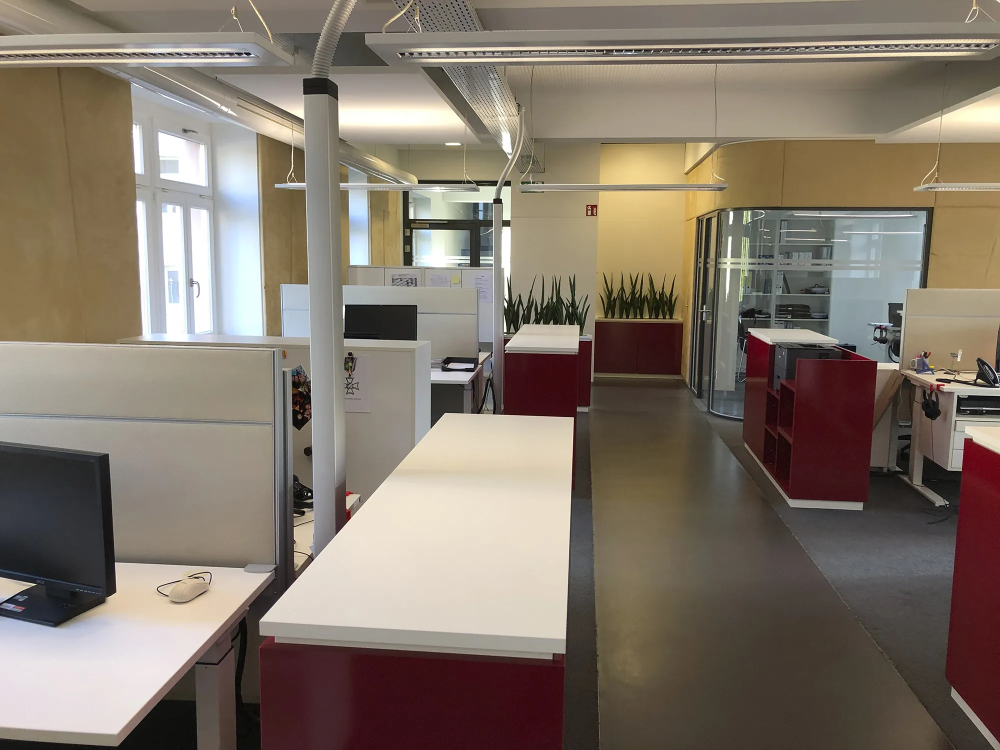 Modernes Großraumbüro mit weißen Schreibtischen, roten Schränken, Monitoren und grünen Pflanzen, beleuchtet durch Tageslicht von großen Fenstern und Deckenleuchten.