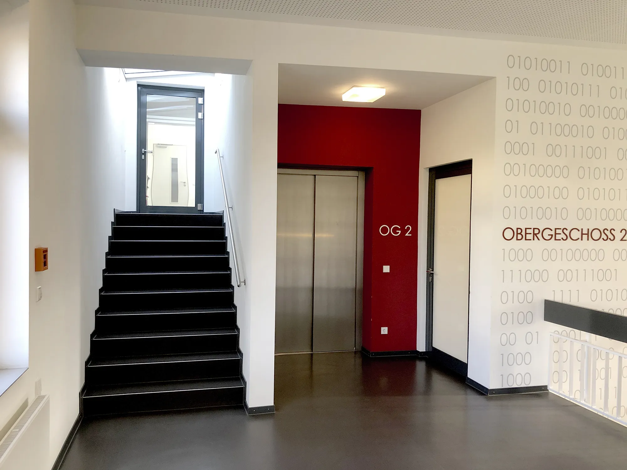 Moderner Gebäudeflur im zweiten Obergeschoss mit schwarzer Treppe zu einer Glastür, Aufzug vor roter Wand mit der Aufschrift „OG 2“ sowie Wandgestaltung mit Binärcode und dem Wort „OBERGESCHOSS 2“.