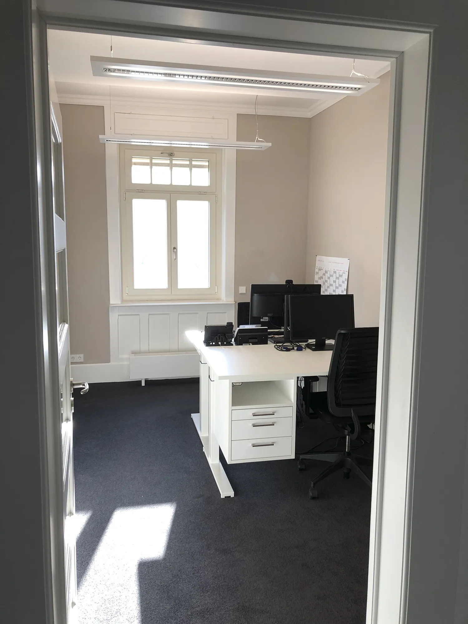 Blick in ein helles Büro mit Teppichboden, weißem Schreibtisch, Bürostuhl und mehreren Monitoren, im Hintergrund ein Fenster mit Tageslichteinfall.