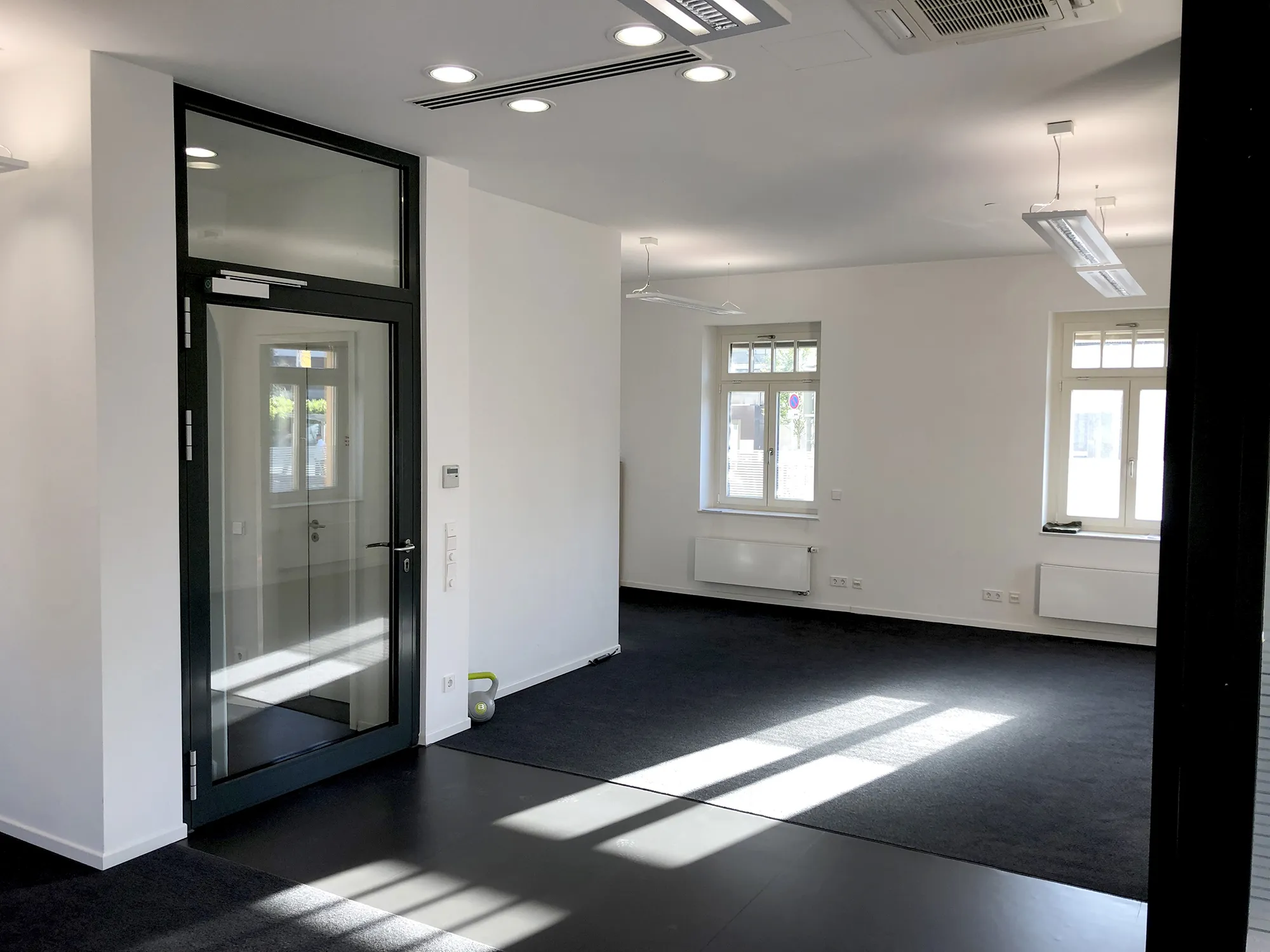 Heller, leerer Büroraum mit weißen Wänden, dunklem Teppichboden und mehreren Fenstern, moderner Glastür mit schwarzem Rahmen sowie Deckenbeleuchtung und Klimaanlage.