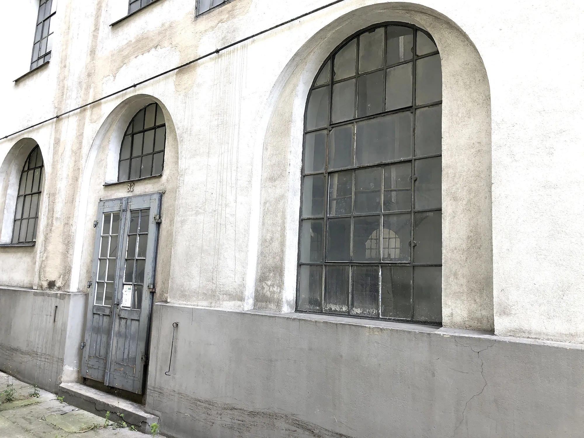 Eingangsbereich eines alten Industriegebäudes mit verwitterter zweiflügeliger Holztür, großen bogenförmigen Sprossenfenstern und heller Fassade.