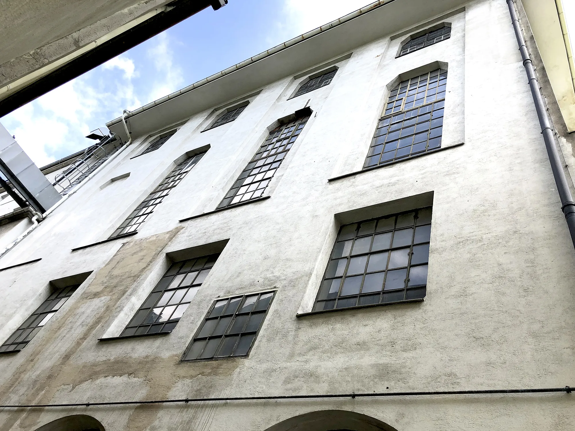 Außenansicht einer mehrstöckigen Industriegebäude-Fassade mit großen Sprossenfenstern und teils verwitterter Putzfläche.