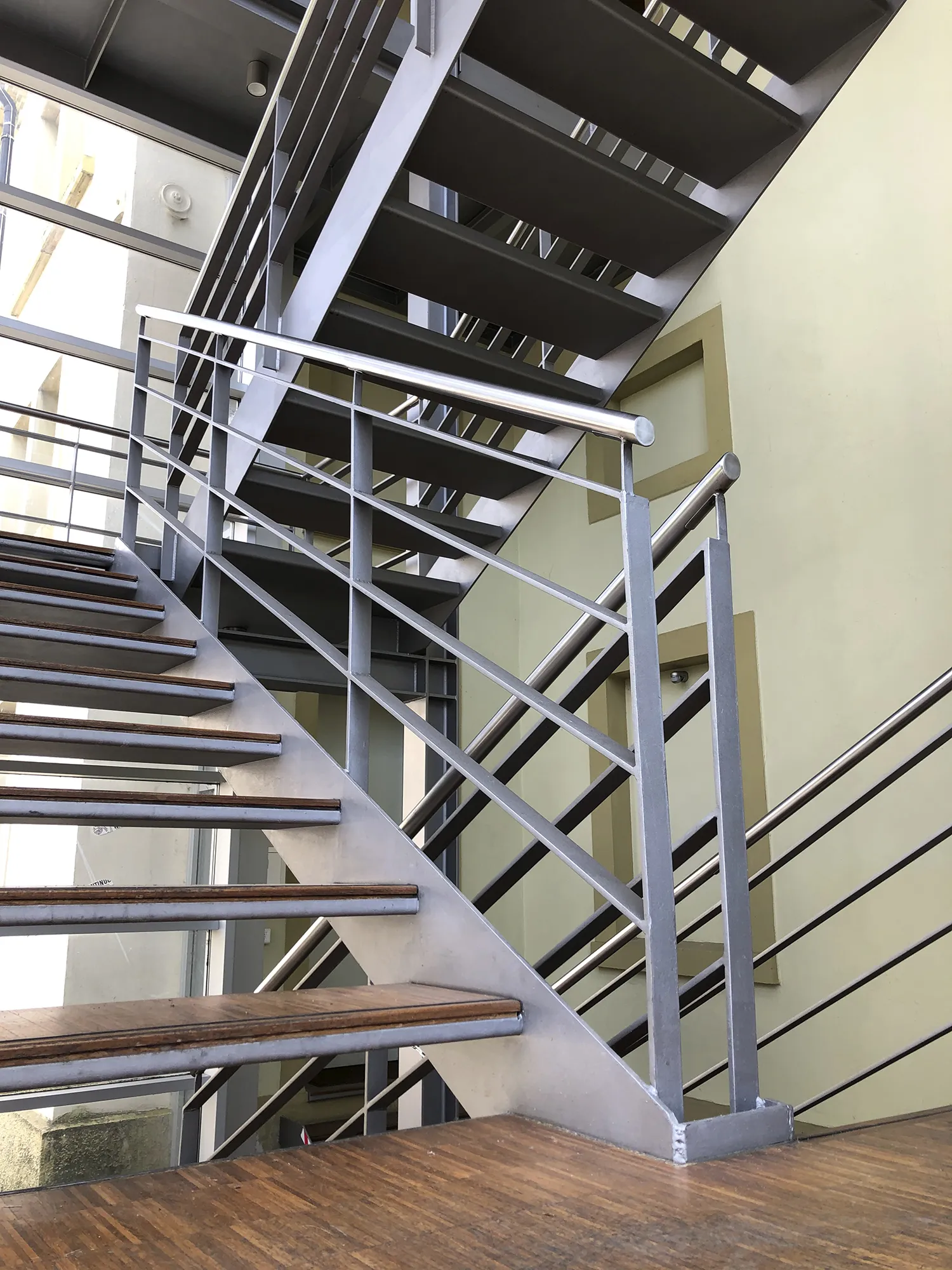Moderne Metalltreppe mit Holzstufen und silbernem Geländer in einem lichtdurchfluteten Treppenhaus mit Glasfassade und helller Wand im Hintergrund.