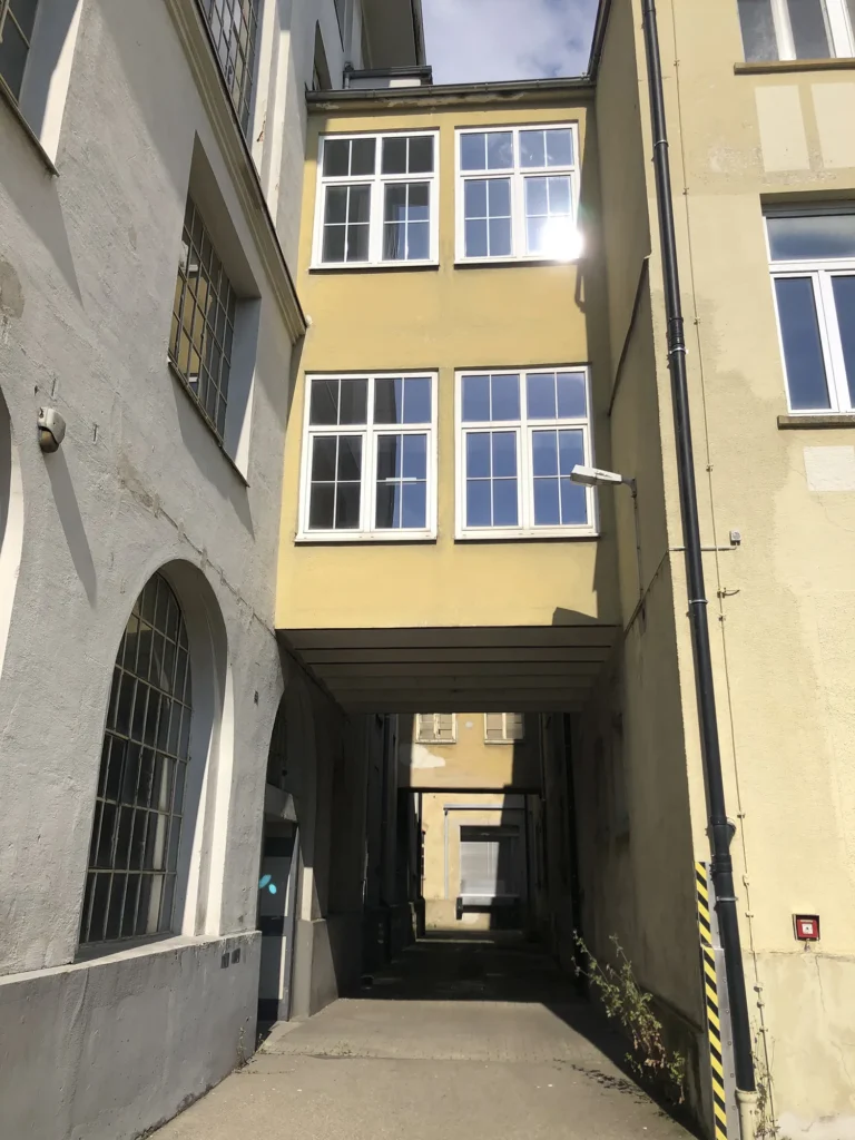 Durchgang zwischen alten Gebäuden mit gelber und grauer Fassade, überbaut mit einem verglasten Übergang im oberen Bereich, Blick durch den schattigen Korridor auf ein weiteres Gebäude im Hintergrund, bei sonnigem Wetter.