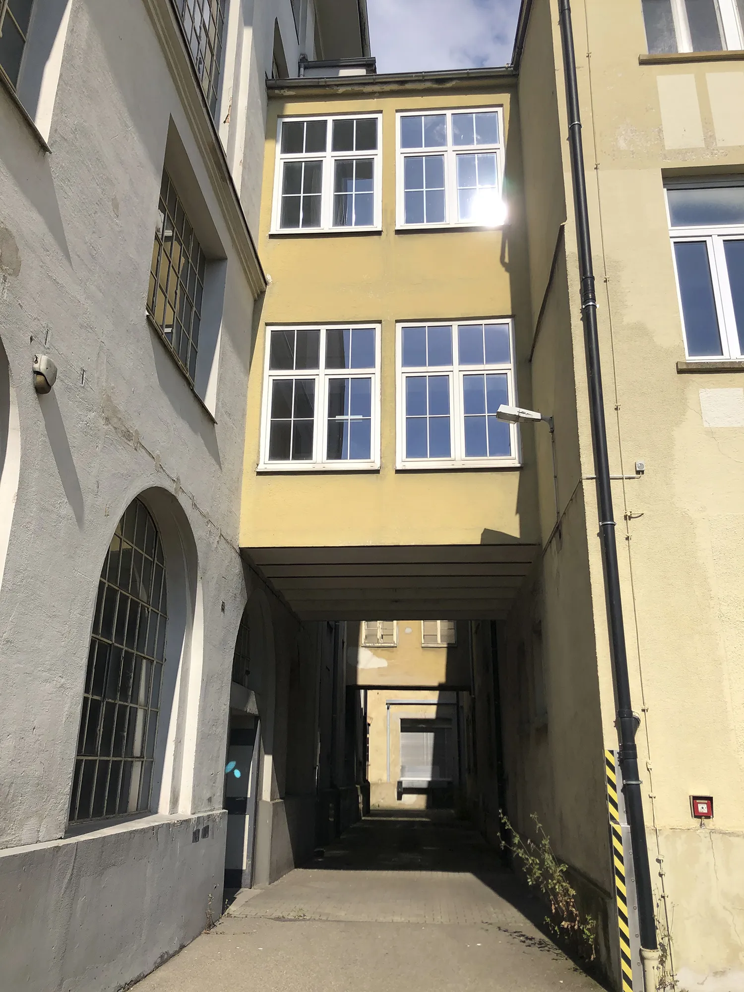 Durchgang zwischen alten Gebäuden mit gelber und grauer Fassade, überbaut mit einem verglasten Übergang im oberen Bereich, Blick durch den schattigen Korridor auf ein weiteres Gebäude im Hintergrund, bei sonnigem Wetter.