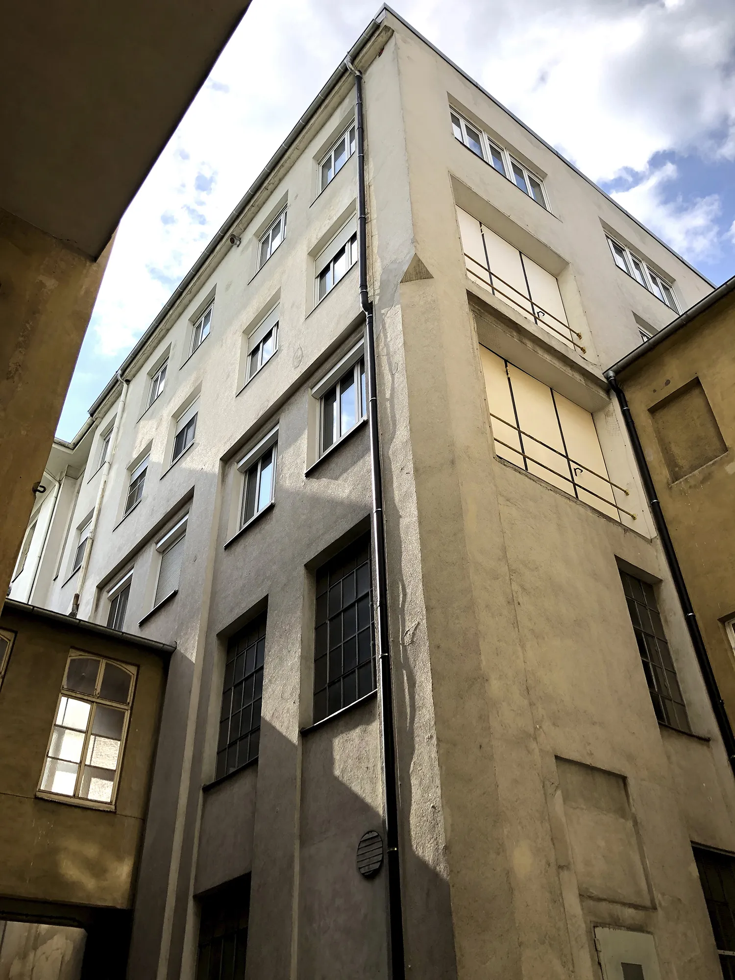 Blick nach oben auf die Fassade eines mehrstöckigen, hell verputzten Gebäudes mit zahlreichen rechteckigen Fenstern, teils mit Rollläden. Die Fassade zeigt Schattenwürfe angrenzender Gebäudeteile, der Himmel ist teils bewölkt.