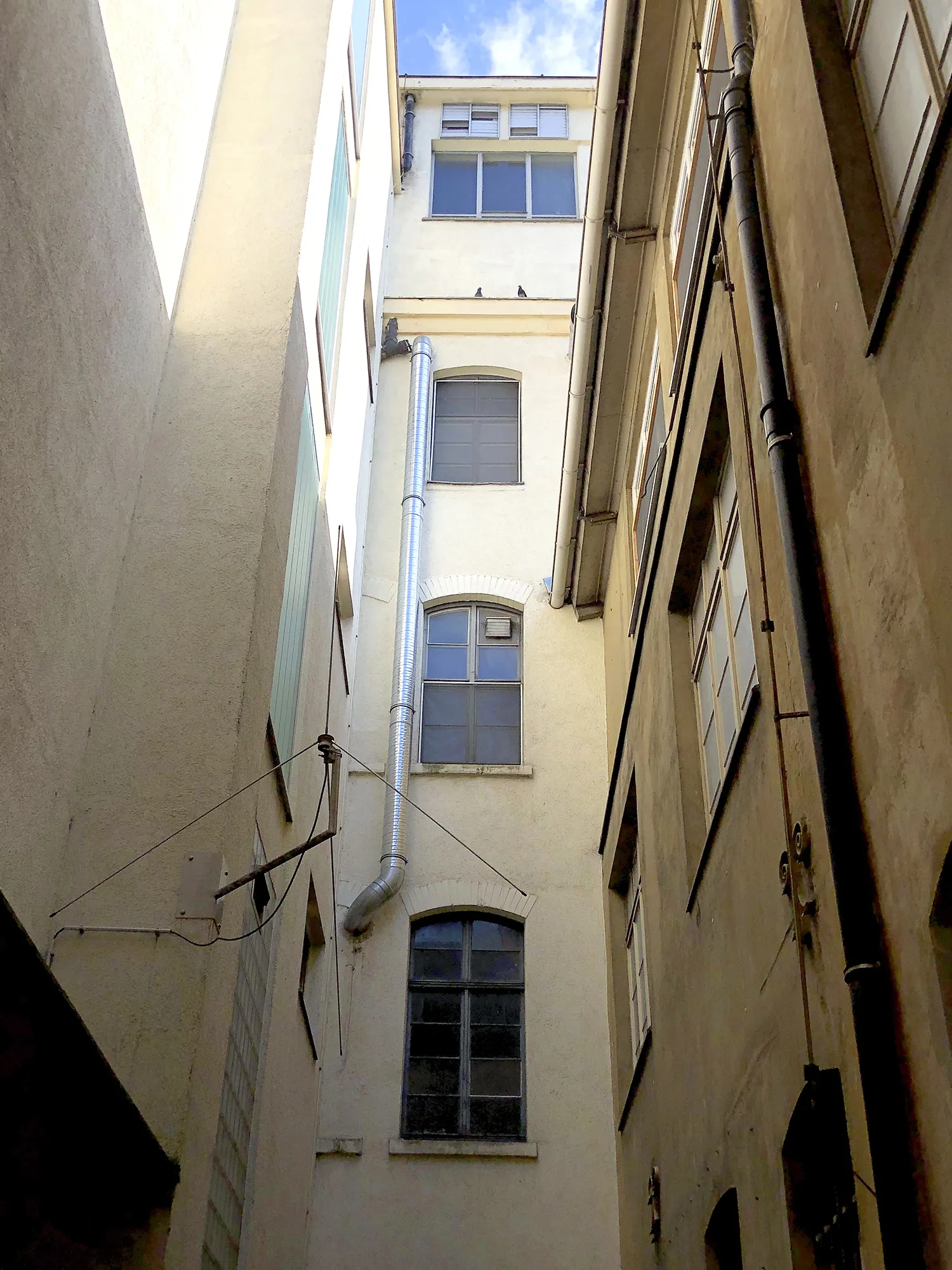 Schmale Gasse zwischen hohen, hellen und beige getönten Gebäuden mit mehreren Fenstern, vertikalen Regenrohren und einem silbernen Abluftrohr, Blick nach oben in Richtung blauen Himmel mit Wolken.