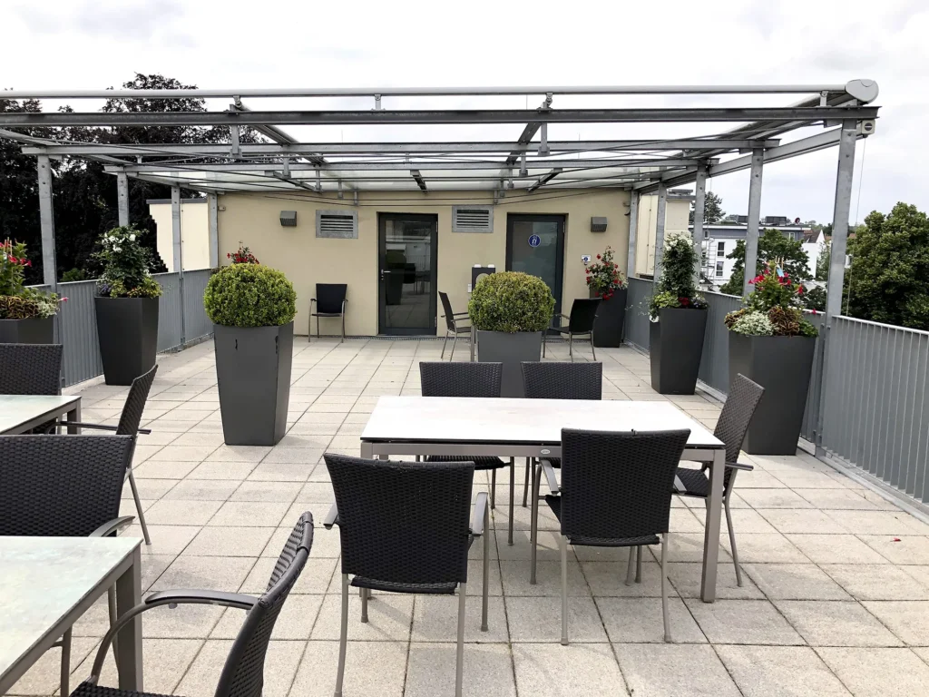 Modern gestaltete Dachterrasse mit Tischen und Stühlen, großen Pflanzkübeln mit verschiedenen Zierpflanzen und einer Überdachung aus Glas und Metall.