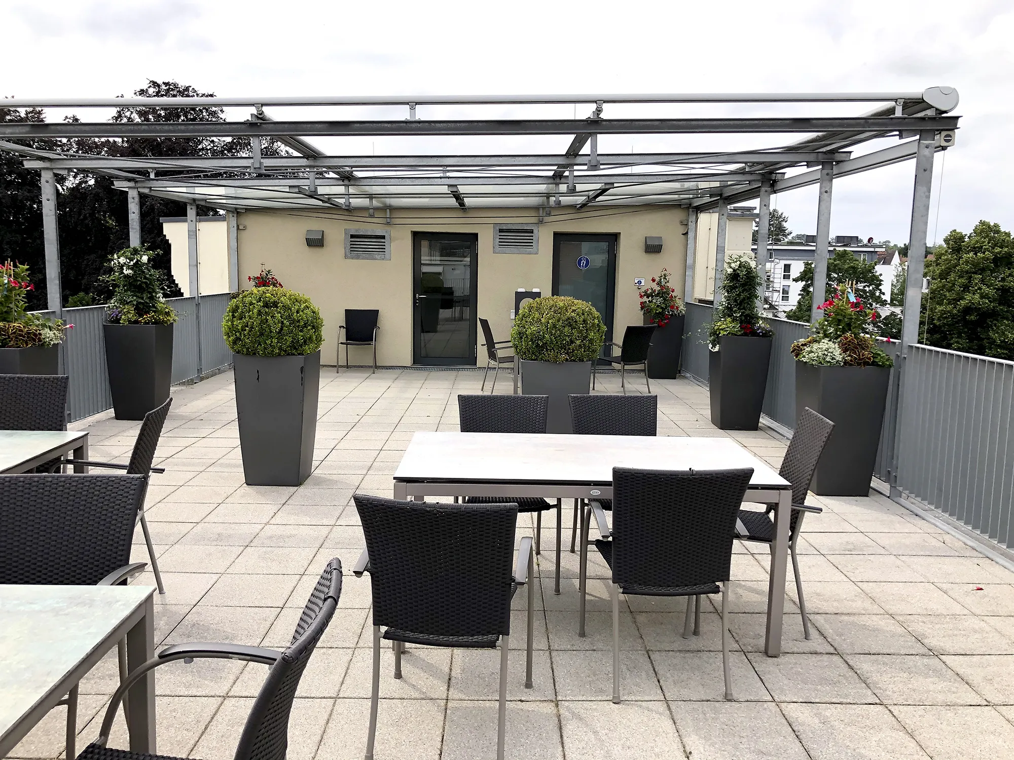 Modern gestaltete Dachterrasse mit Tischen und Stühlen, großen Pflanzkübeln mit verschiedenen Zierpflanzen und einer Überdachung aus Glas und Metall.