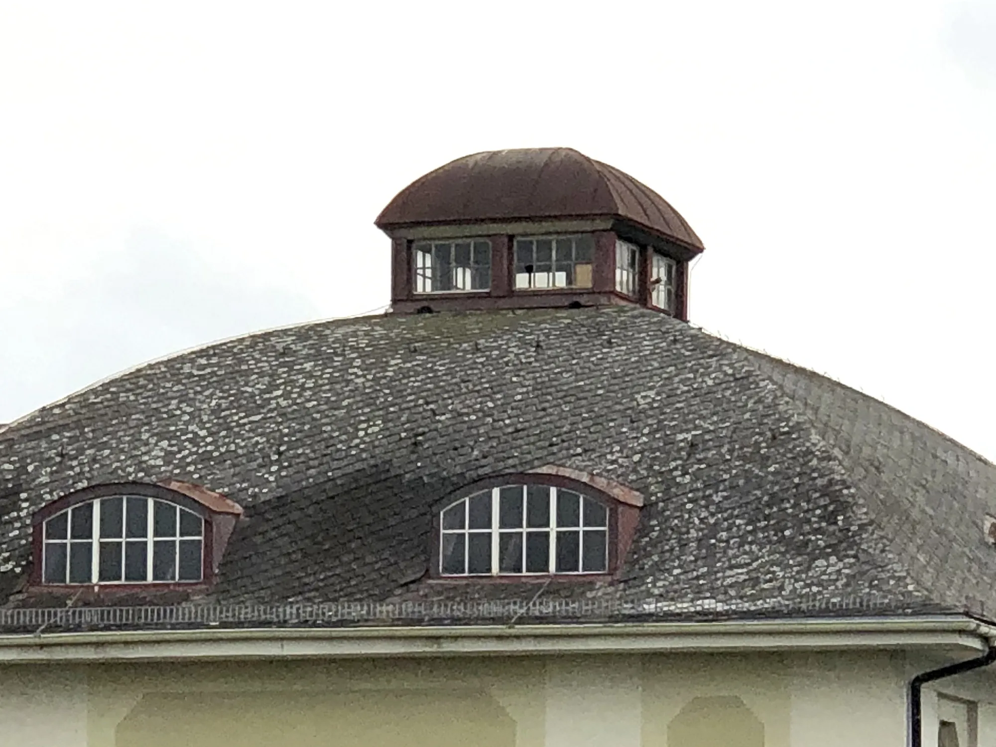 Nahaufnahme eines gewölbten Schieferdachs mit zwei Dachgauben und einem kleinen Dachaufbau mit roten Fensterrahmen, vor grauem Himmel.