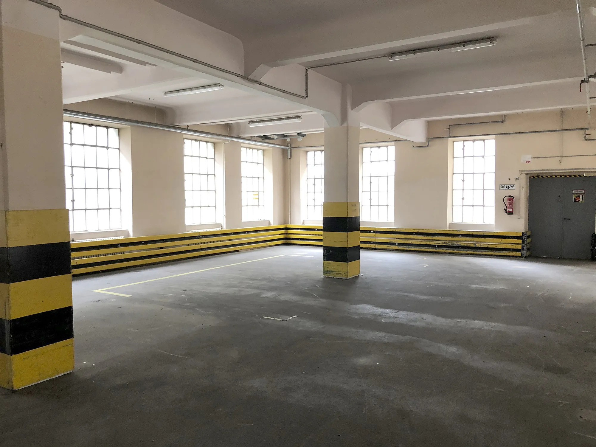 Leere Industriehalle mit großen Fenstern, gelb-schwarz markierten Pfeilern und Wänden sowie einem Betonboden.