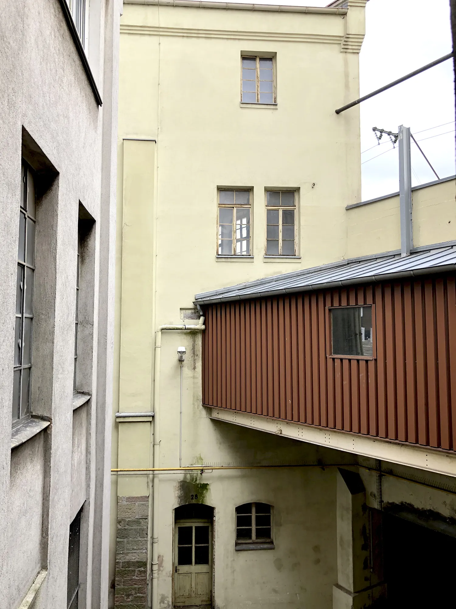 Innenhofansicht eines Industriegebäudes mit gelber Fassade, mehreren Fenstern, einer roten geschlossenen Übergangsbrücke und einer alten Holztür im Erdgeschoss.