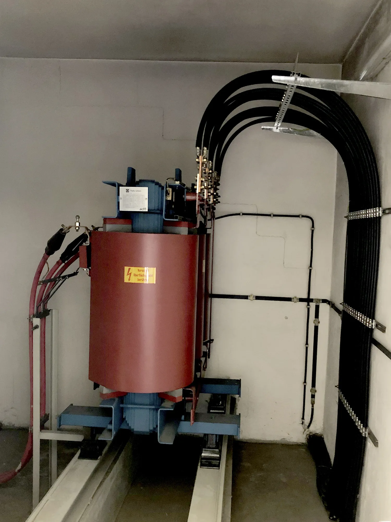 Elektrischer Transformator mit rotem Gehäuse, angeschlossenen Hochspannungskabeln und montiert auf einem blauen Metallrahmen in einem Technikraum.