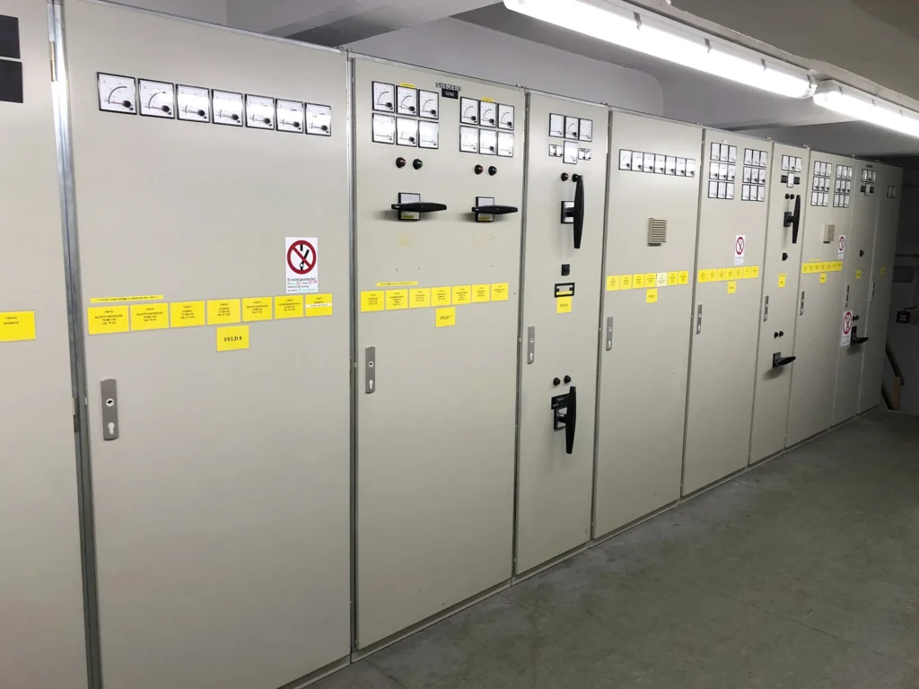Reihe von einer großen, beige-farbenen Schaltanlage mit analogen Messinstrumenten, Schaltern und gelben Beschriftungsschildern in einem Technikraum.