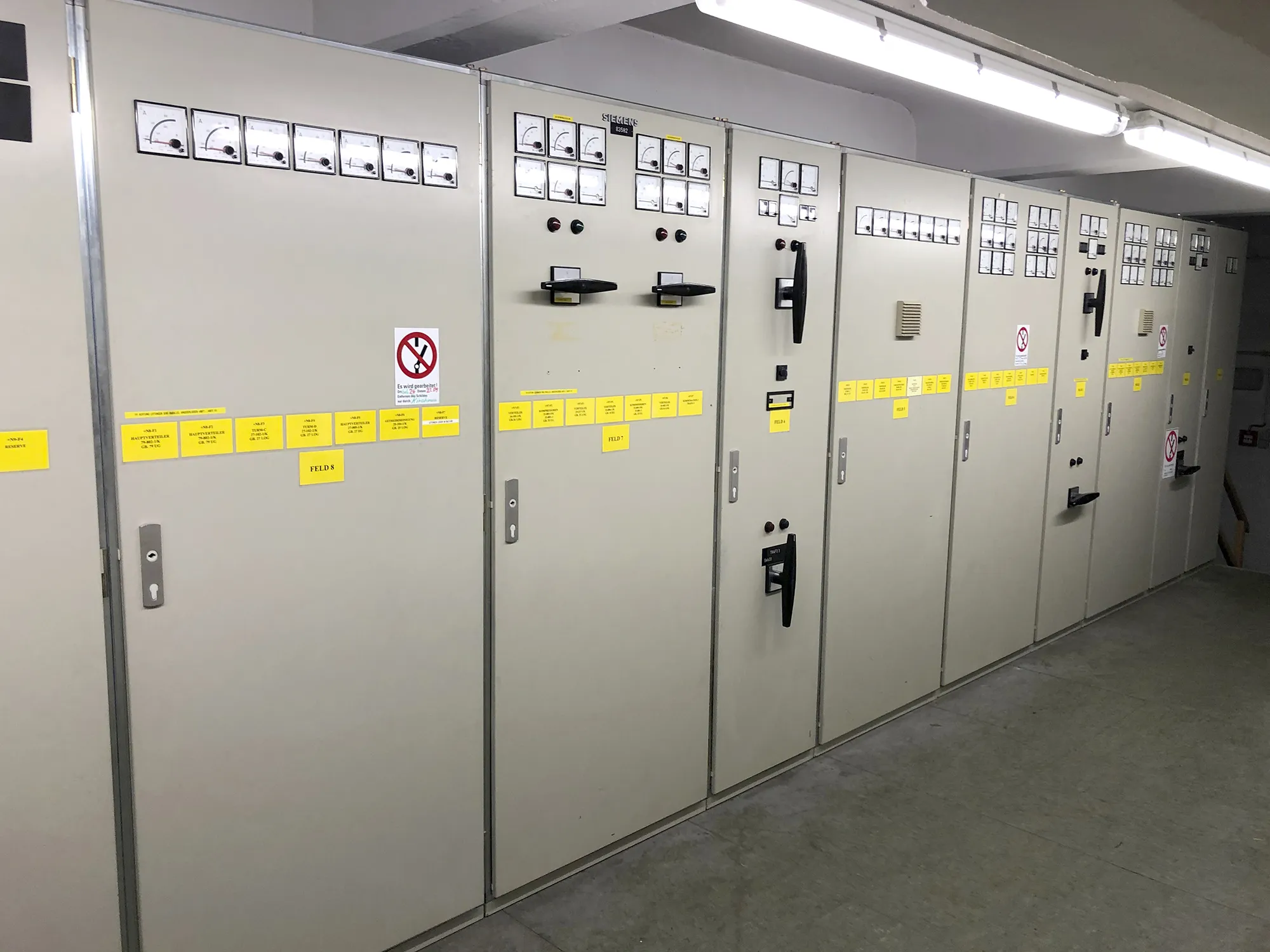 Reihe von einer großen, beige-farbenen Schaltanlage mit analogen Messinstrumenten, Schaltern und gelben Beschriftungsschildern in einem Technikraum.