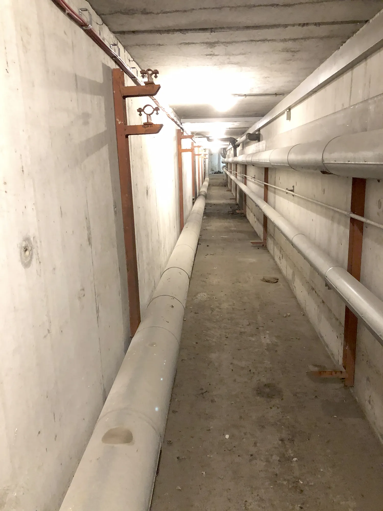 Langer, schmaler Versorgungsgang mit freiliegenden Rohrleitungen an beiden Wänden und Betonboden, beleuchtet durch Deckenlampen.