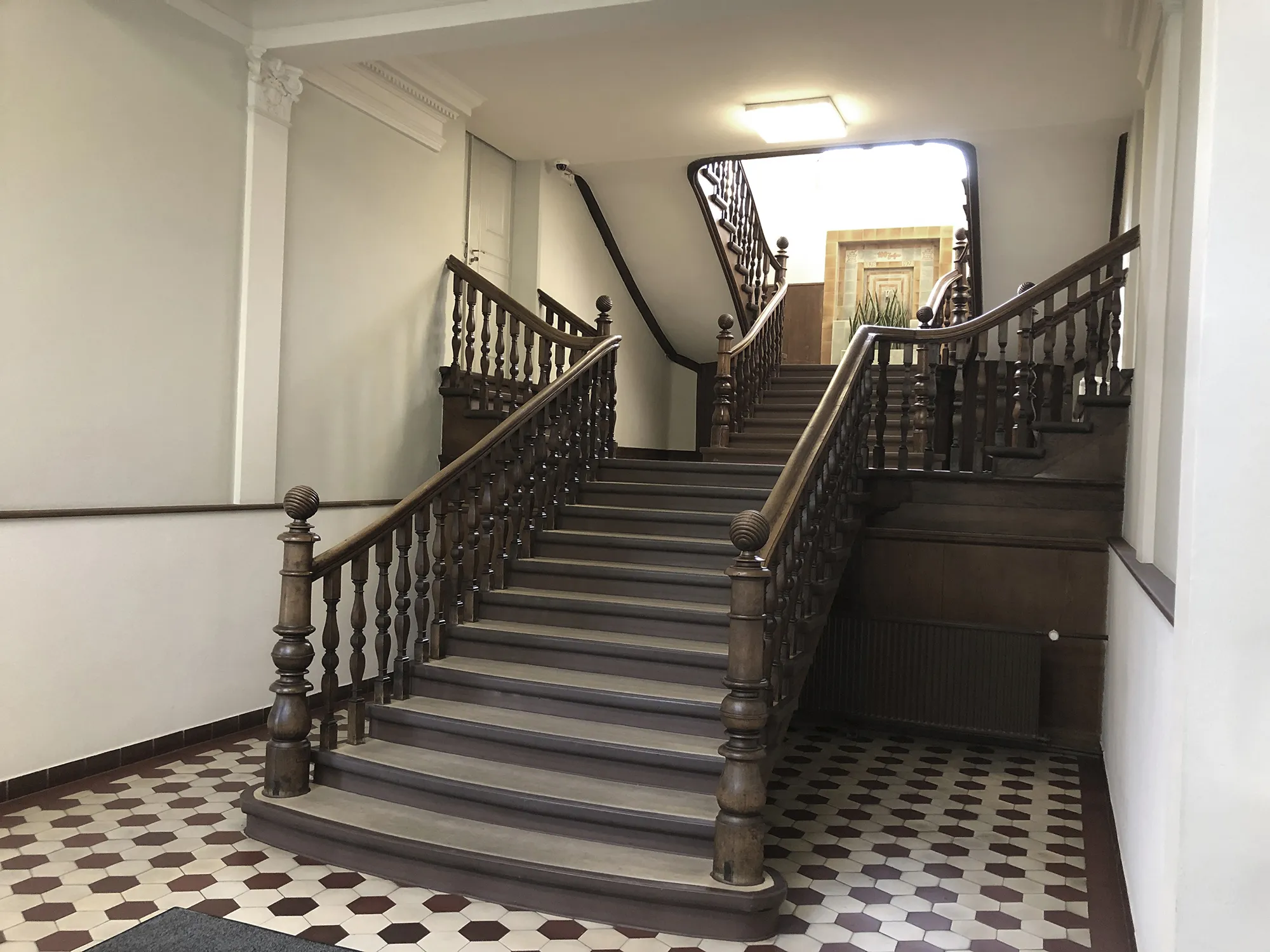 Breite Holztreppe mit verzierten Geländern und Balustern in einem Eingangsbereich mit rot-weiß gemustertem Fliesenboden und heller Wandgestaltung.