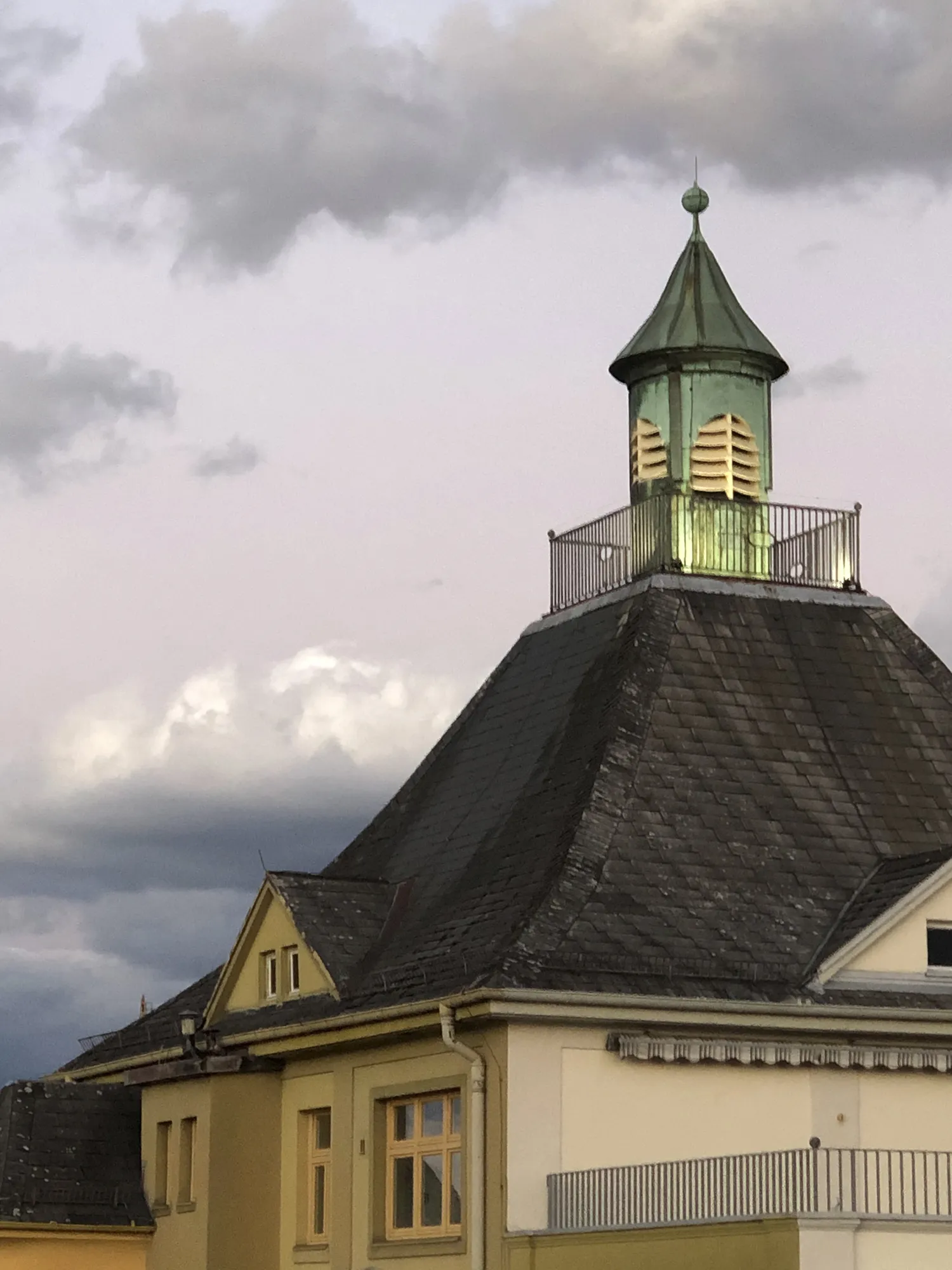 Turmspitze eines historischen Gebäudes mit grün oxidiertem Kupferdach, umlaufendem Geländer und dunklem Schieferdach darunter, vor bewölktem Himmel in der Abenddämmerung.