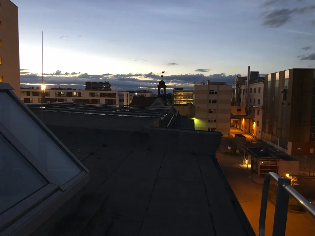 Blick vom Dach auf Ludwigsburg bei Sonnenuntergang, mit Gebäuden im Vordergrund, einer Silhouette eines Turms in der Mitte und einem wolkenverhangenen Himmel im Hintergrund.