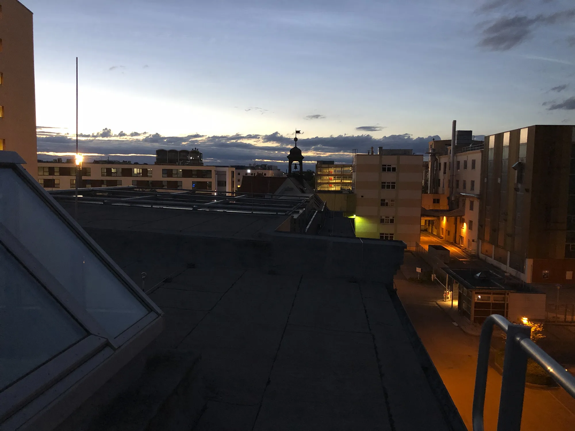 Blick vom Dach auf Ludwigsburg bei Sonnenuntergang, mit Gebäuden im Vordergrund, einer Silhouette eines Turms in der Mitte und einem wolkenverhangenen Himmel im Hintergrund.