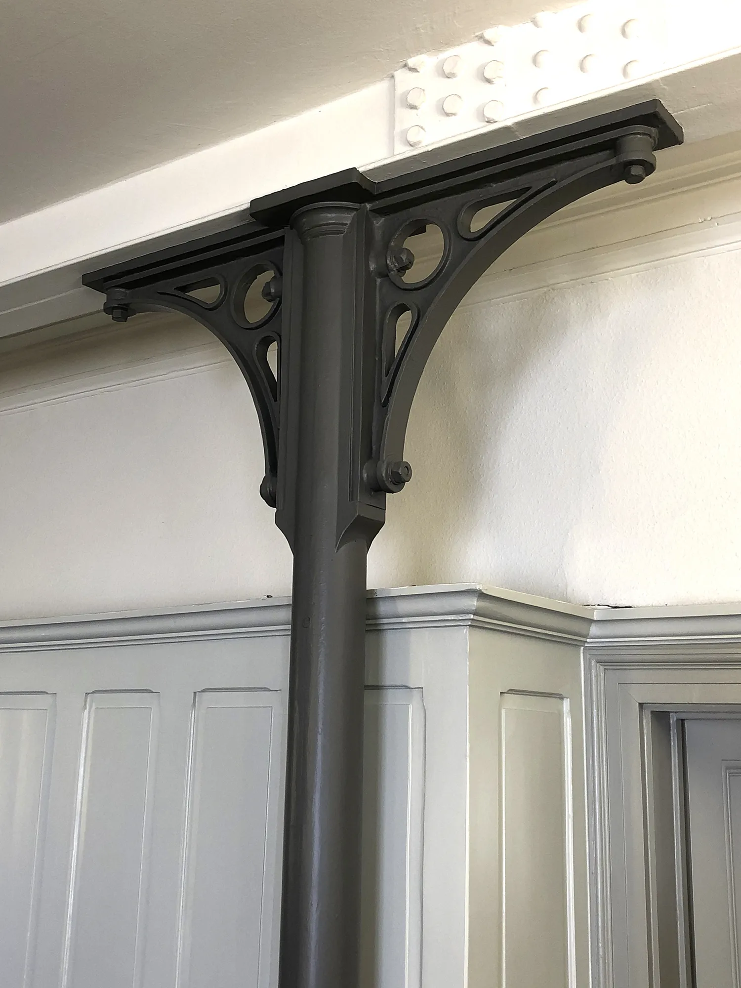 Schwarze Stahlstütze mit seitlichen Zierverstrebungen in Dreiecksform, montiert unter einer weißen Decke neben einer grauen Wandverkleidung.