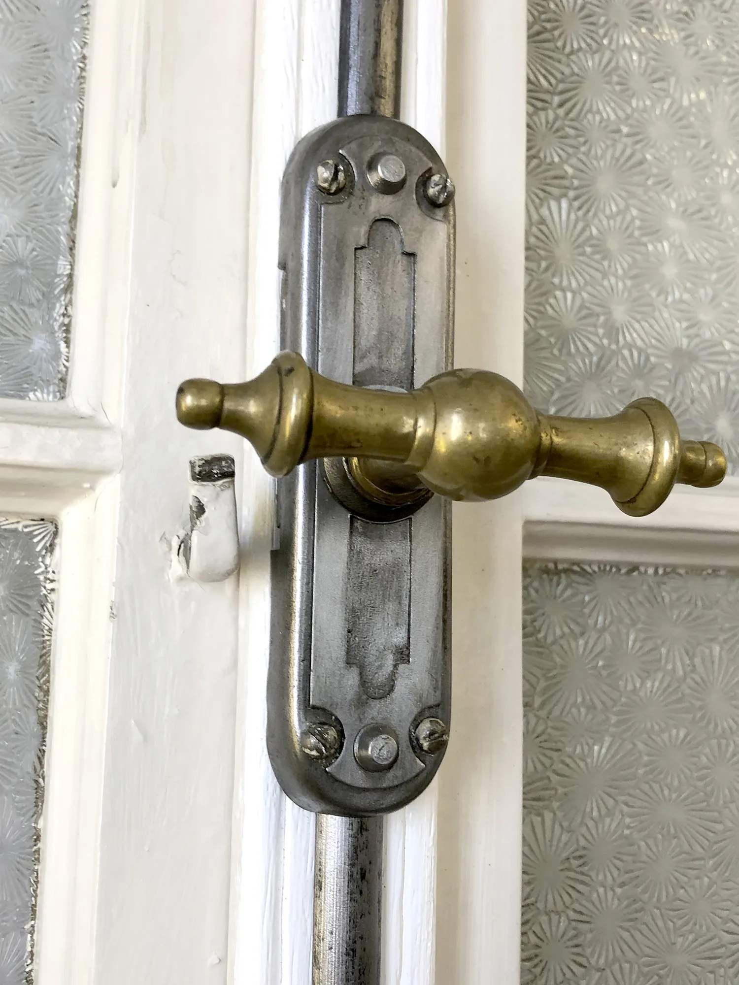 Detailaufnahme eines Türgriffs aus Messing mit verzierter Metallplatte, montiert an einer weißen Holztür mit strukturiertem Glas.