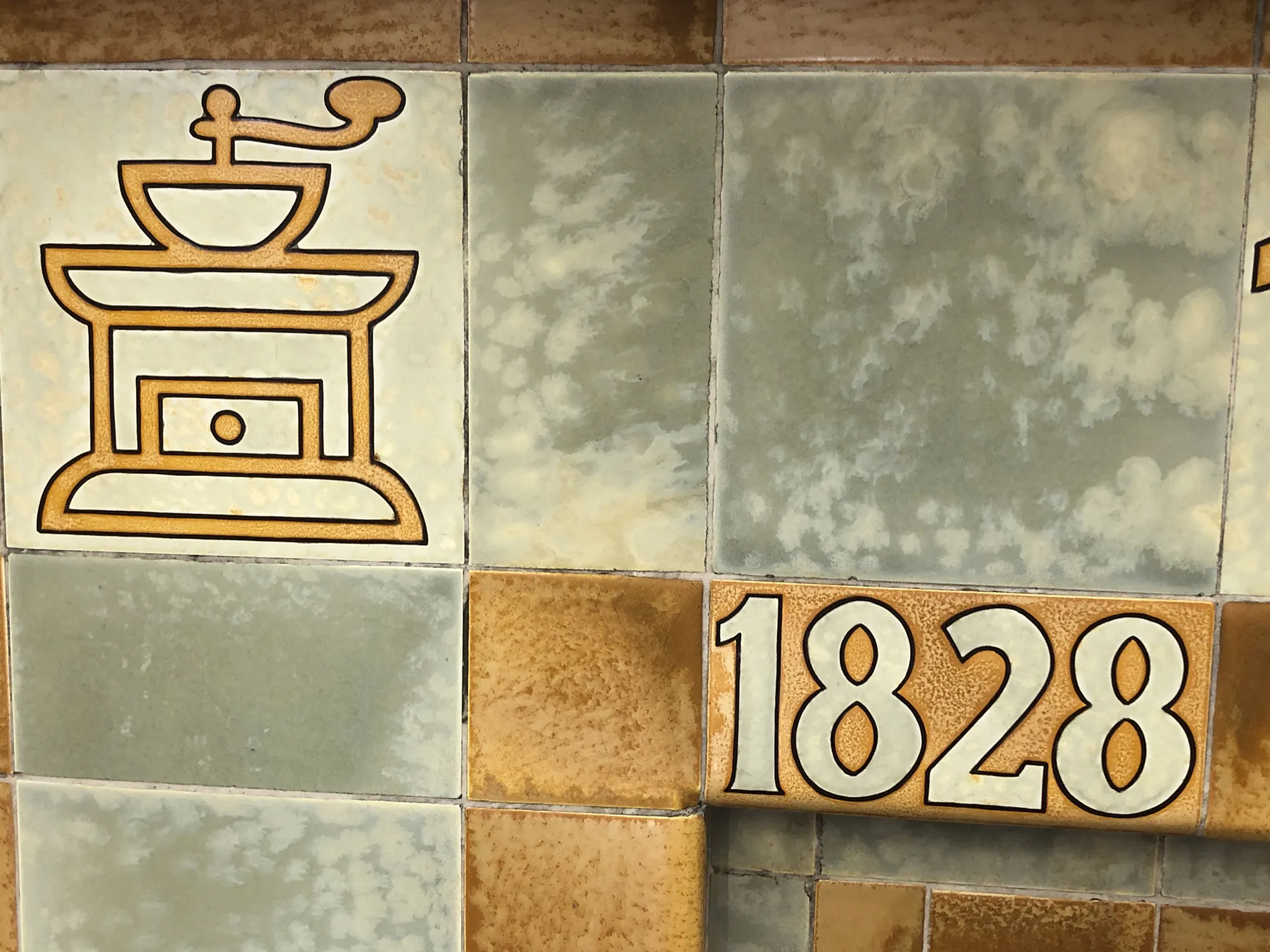 Keramikkacheln in Grün- und Brauntönen, links mit einer stilisierten Darstellung einer Kaffeemühle in brauner Linienführung, rechts unten mit der Zahl „1828“ in großen weißen Ziffern auf braun gemustertem Hintergrund.