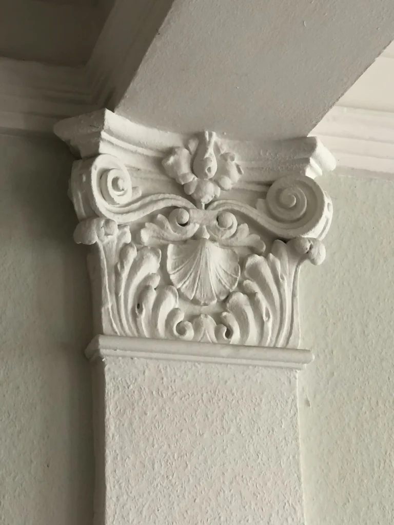 Detailaufnahme eines weißen, verzierten Kapitells mit floralen Ornamenten, Voluten und einer zentralen Muschelform.