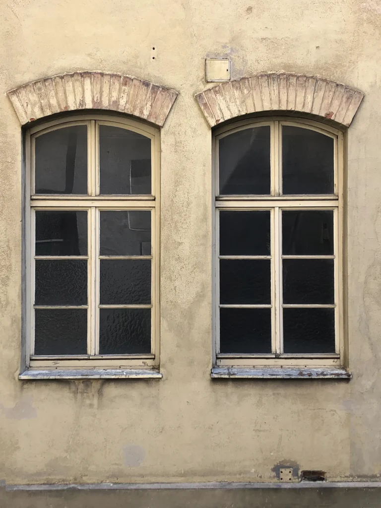 Zwei nebeneinanderliegende, hohe Holzfenster mit Rundbogen und Sprossen, eingefasst in eine verwitterte Putzfassade mit gemauerten Bogenstürzen.