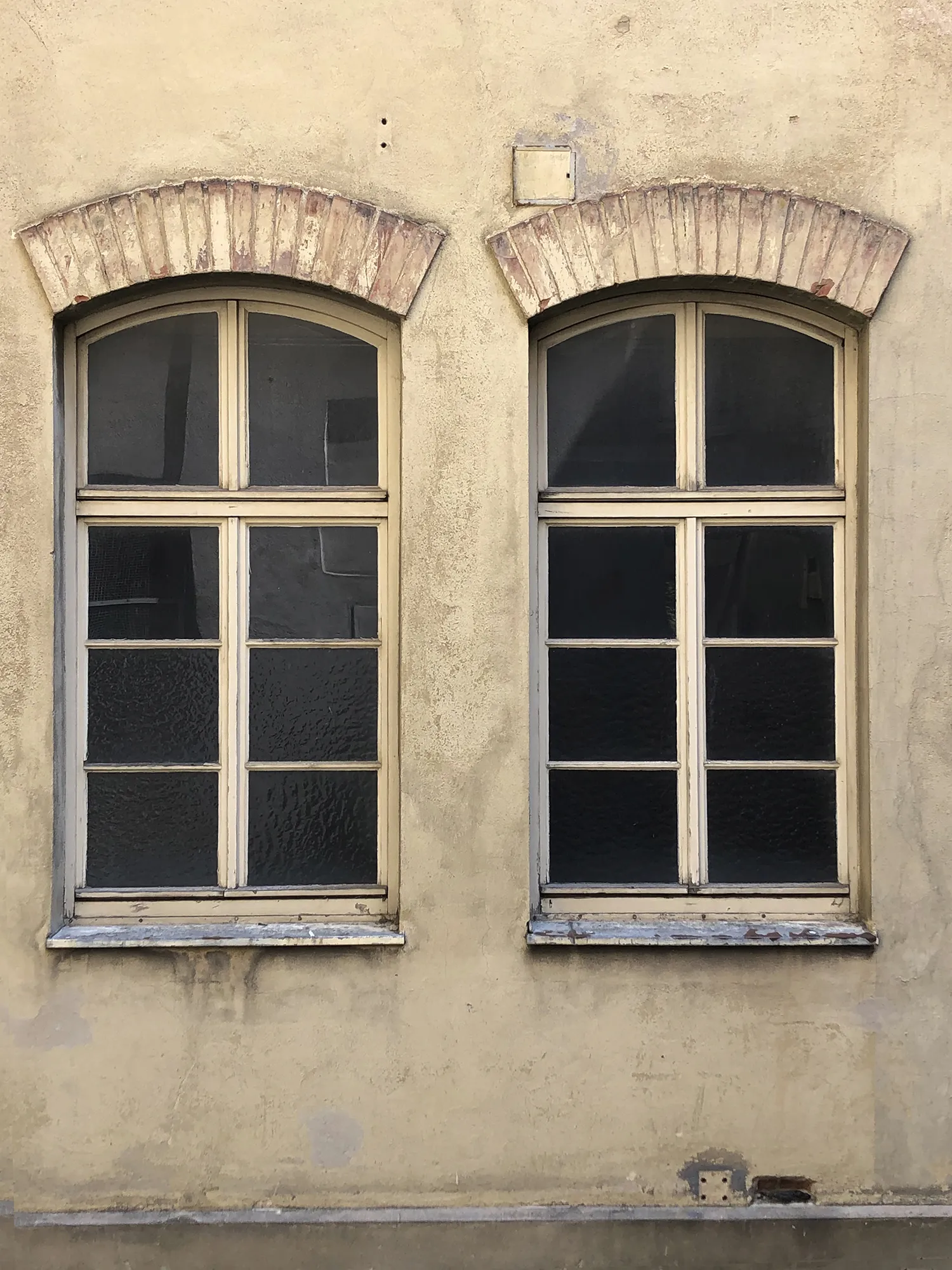 Zwei nebeneinanderliegende, hohe Holzfenster mit Rundbogen und Sprossen, eingefasst in eine verwitterte Putzfassade mit gemauerten Bogenstürzen.
