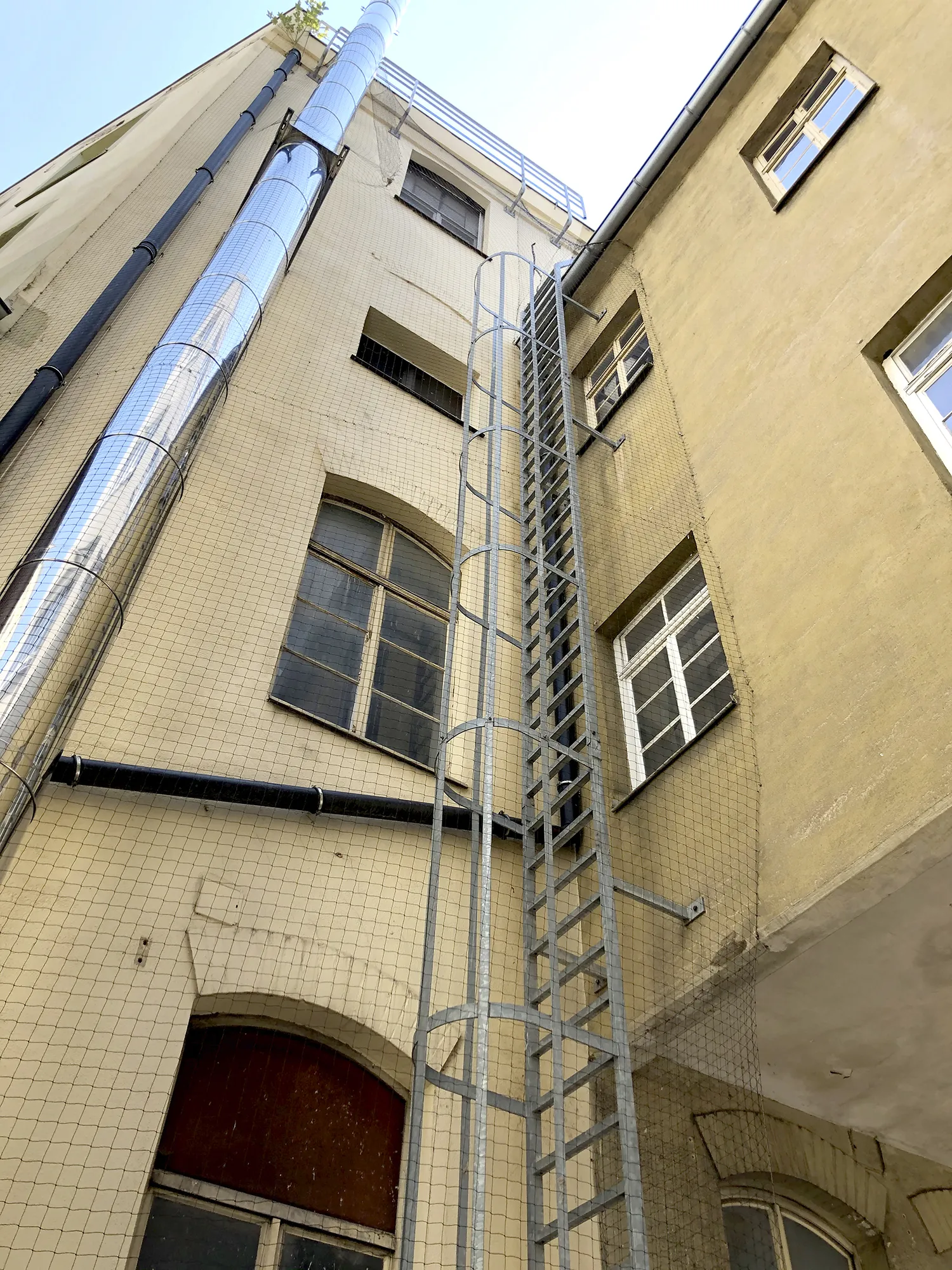 Blick nach oben auf den Innenhof eines Industriegebäudes mit Metallaußenleiter und silbernen Rohrleitungen, gesichert durch ein umlaufendes Schutznetz.