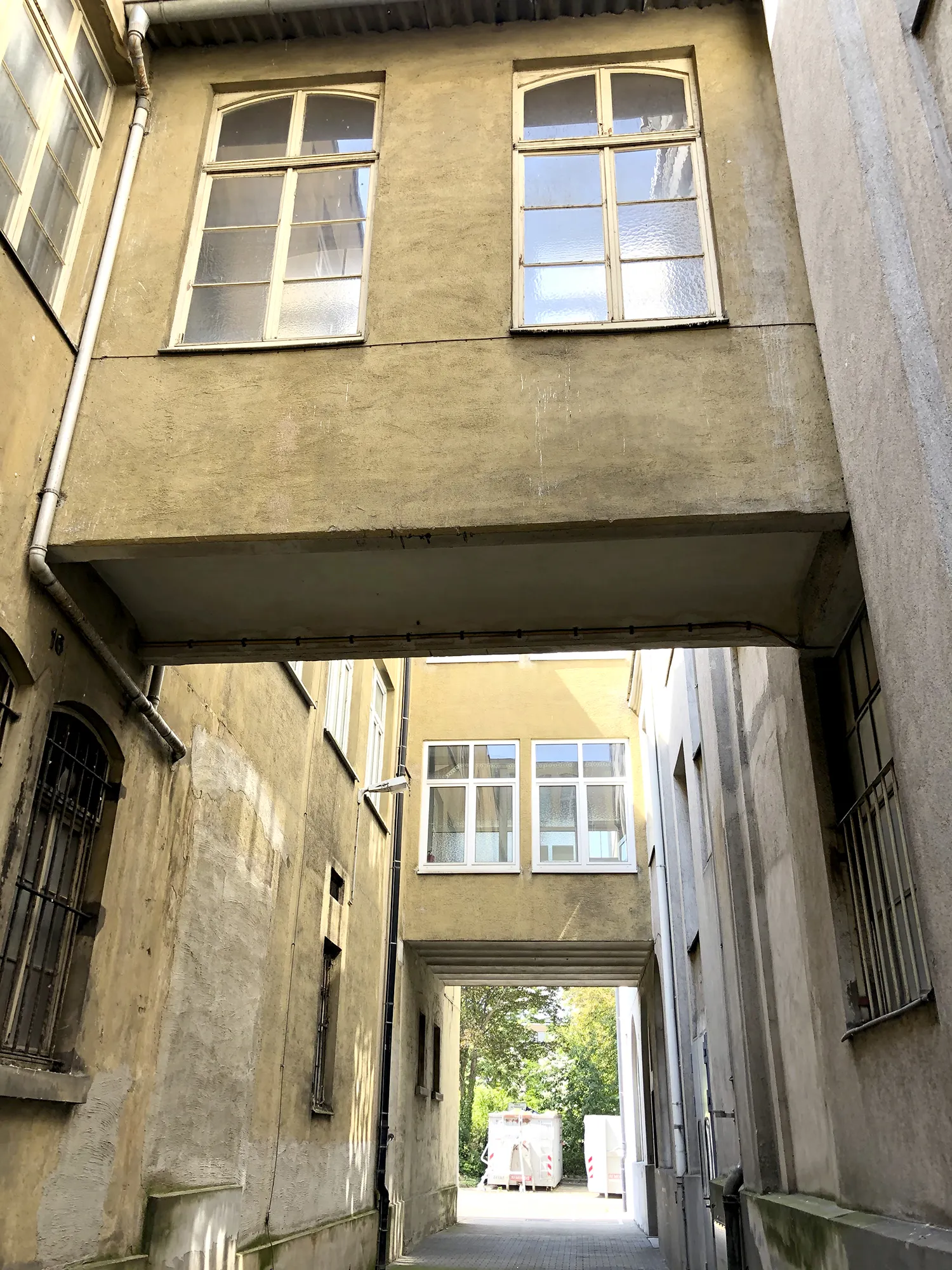 Schmaler Durchgang zwischen alten Gebäuden mit zwei verglasten Übergängen im oberen Bereich, Blick hinaus auf einen sonnigen Hof mit Containern und Bäumen im Hintergrund.