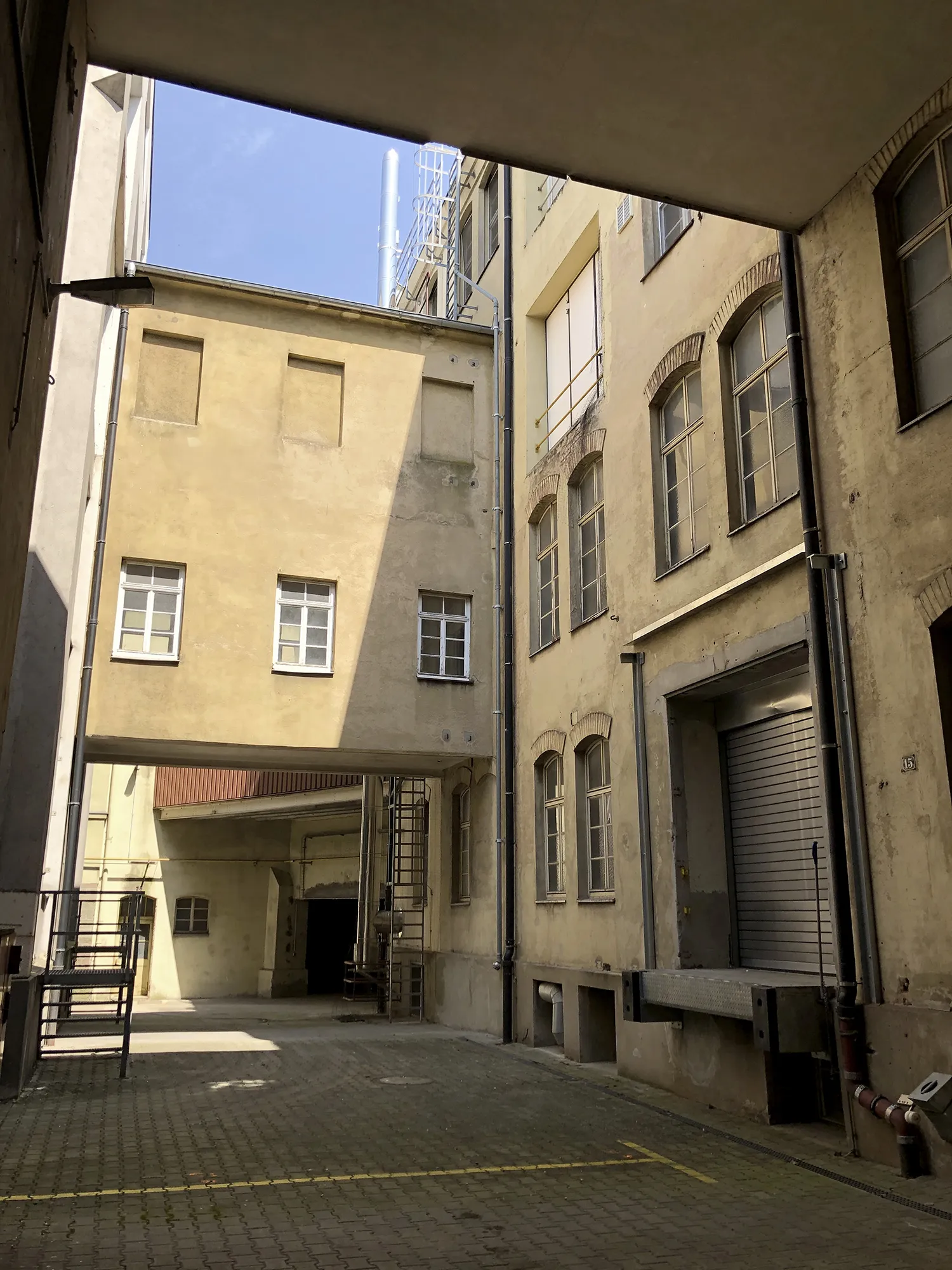 Innenhof des Caro Carrés mit gelblich-beigen Fassaden, mehreren Fenstern, einer Verbindungsbrücke zwischen den Gebäuden und einem Rolltor auf der rechten Seite.