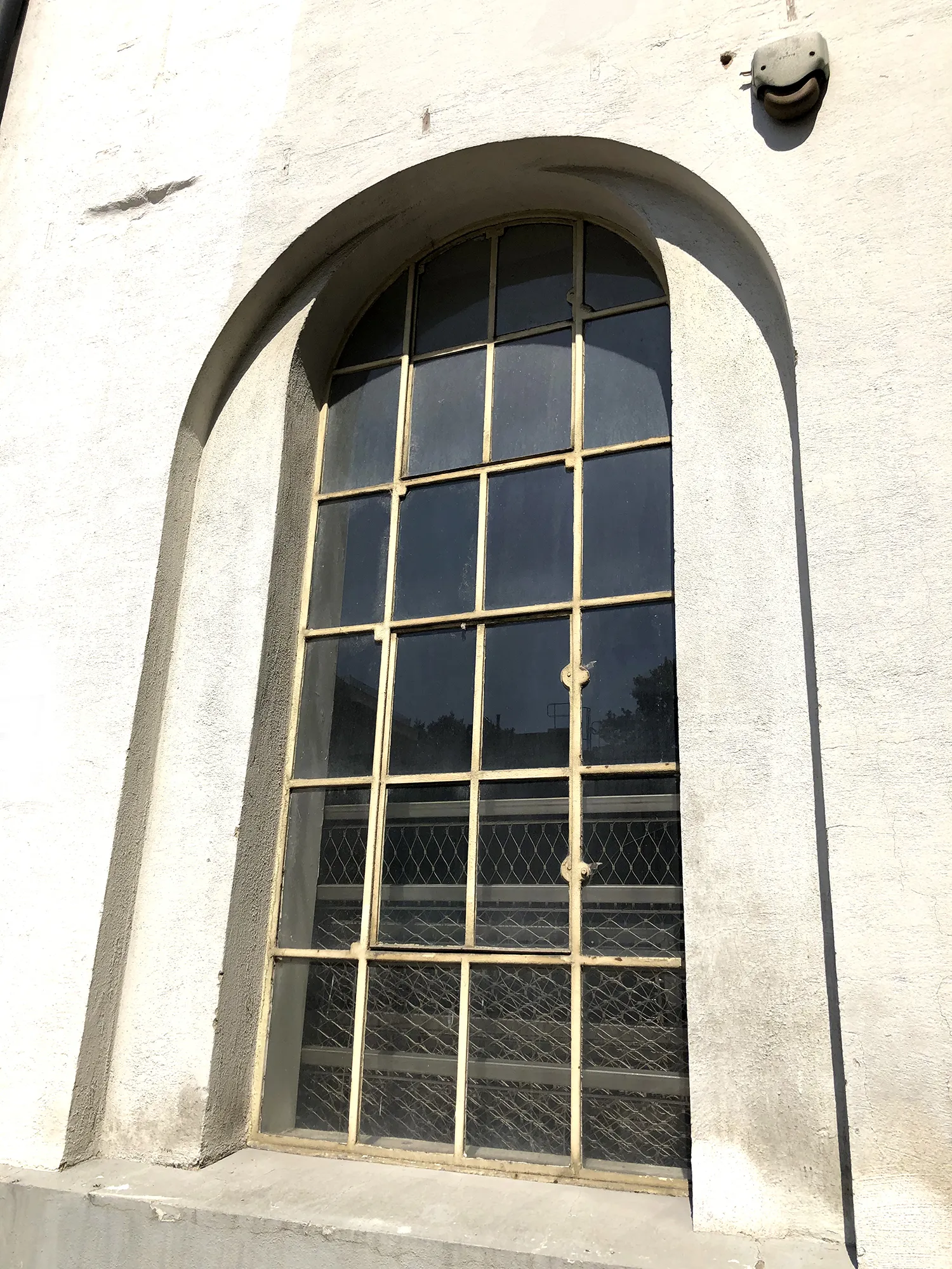 Hoher, schmaler Bogenfensterrahmen aus Metall mit mehreren kleinen Scheiben in einer weißen Putzfassade, im Sonnenlicht.
