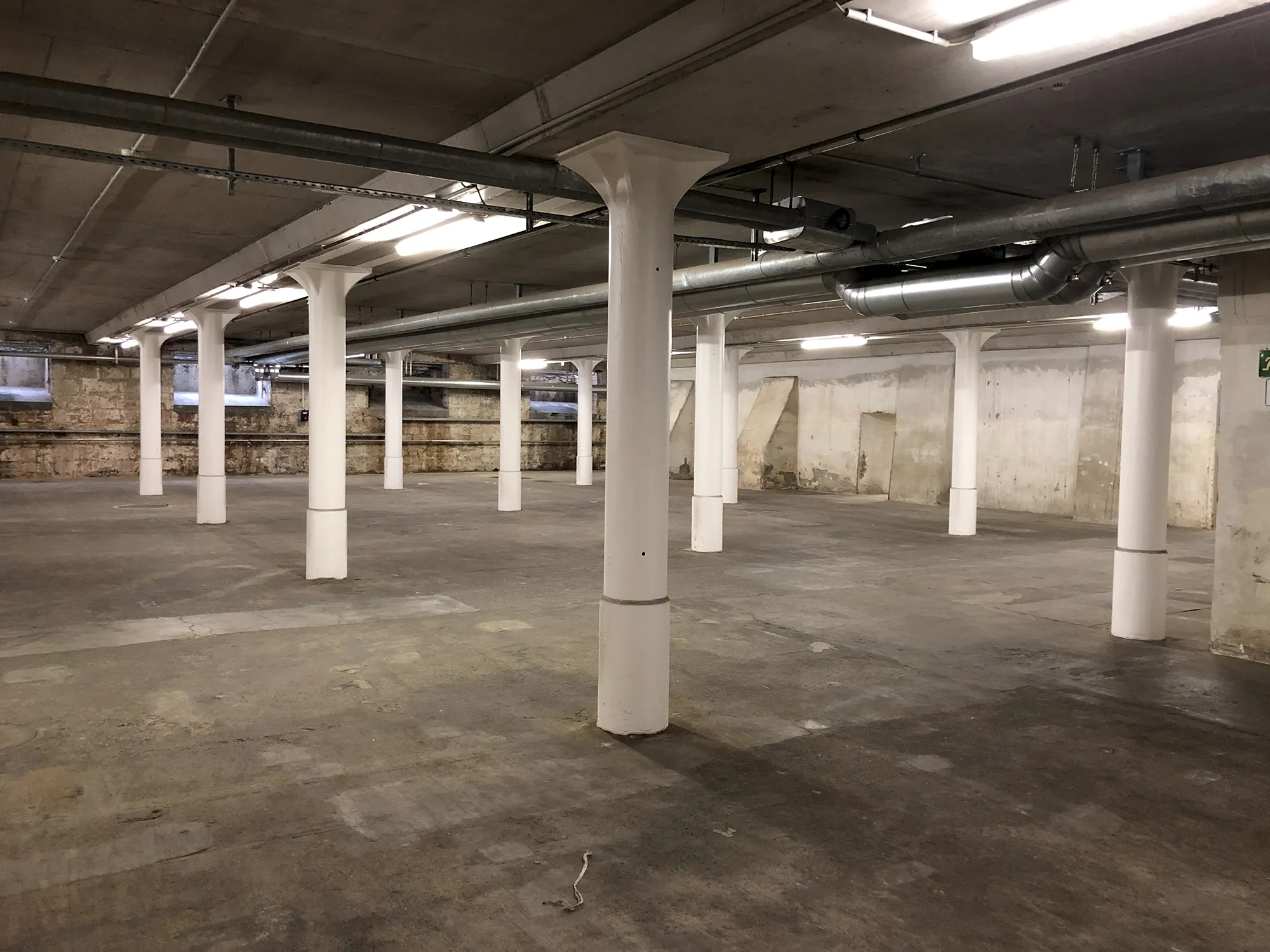 Großer, leerer Kellerraum mit weißen Stützsäulen, Betonboden, alten Mauerwänden und freiliegenden Lüftungsrohren unter der Decke.