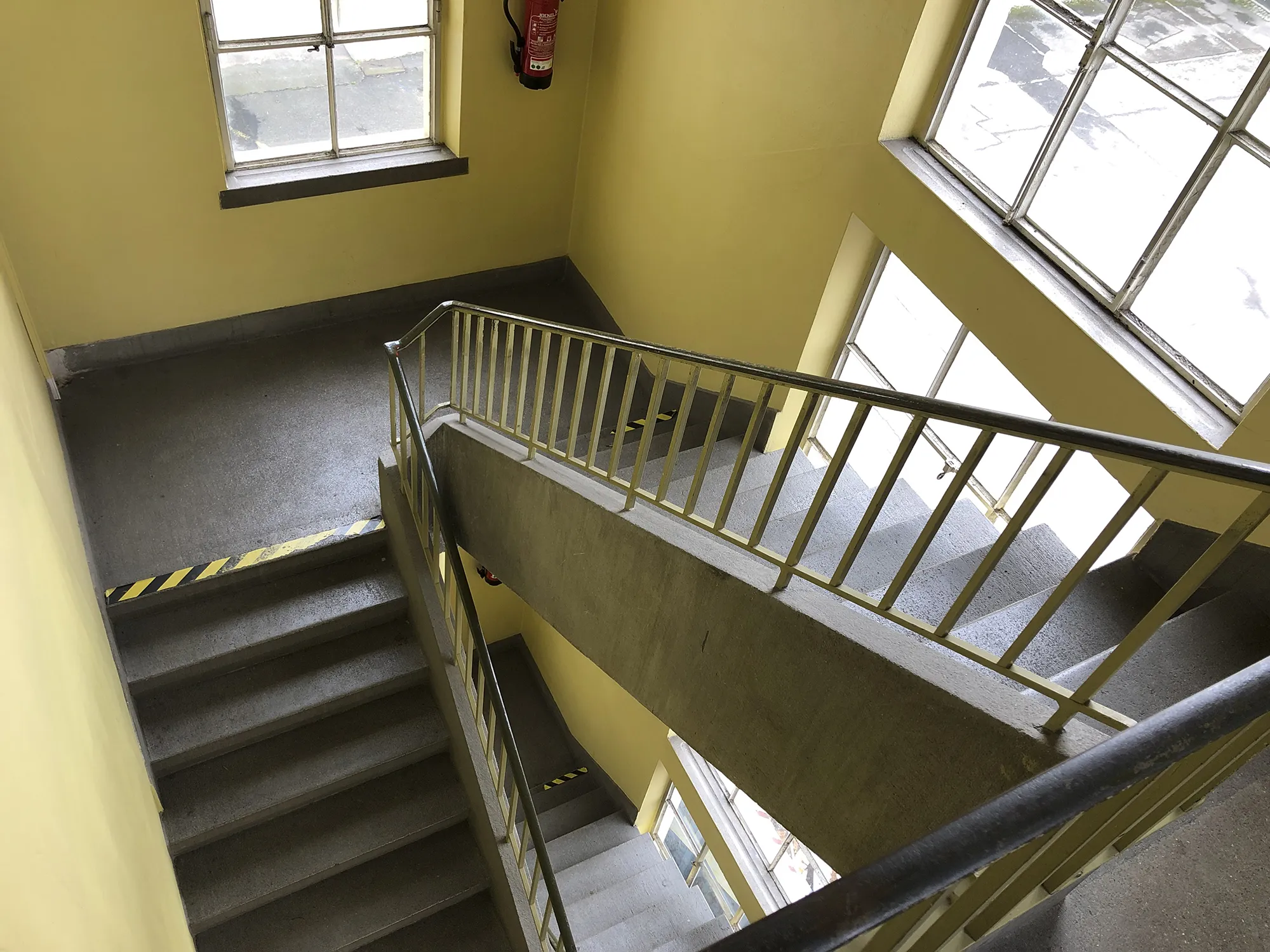Treppenhaus mit grauen Stufen, gelben Wänden, großen Fenstern und Metallgeländer, markiert mit gelb-schwarzen Sicherheitsstreifen an den Stufenkanten.