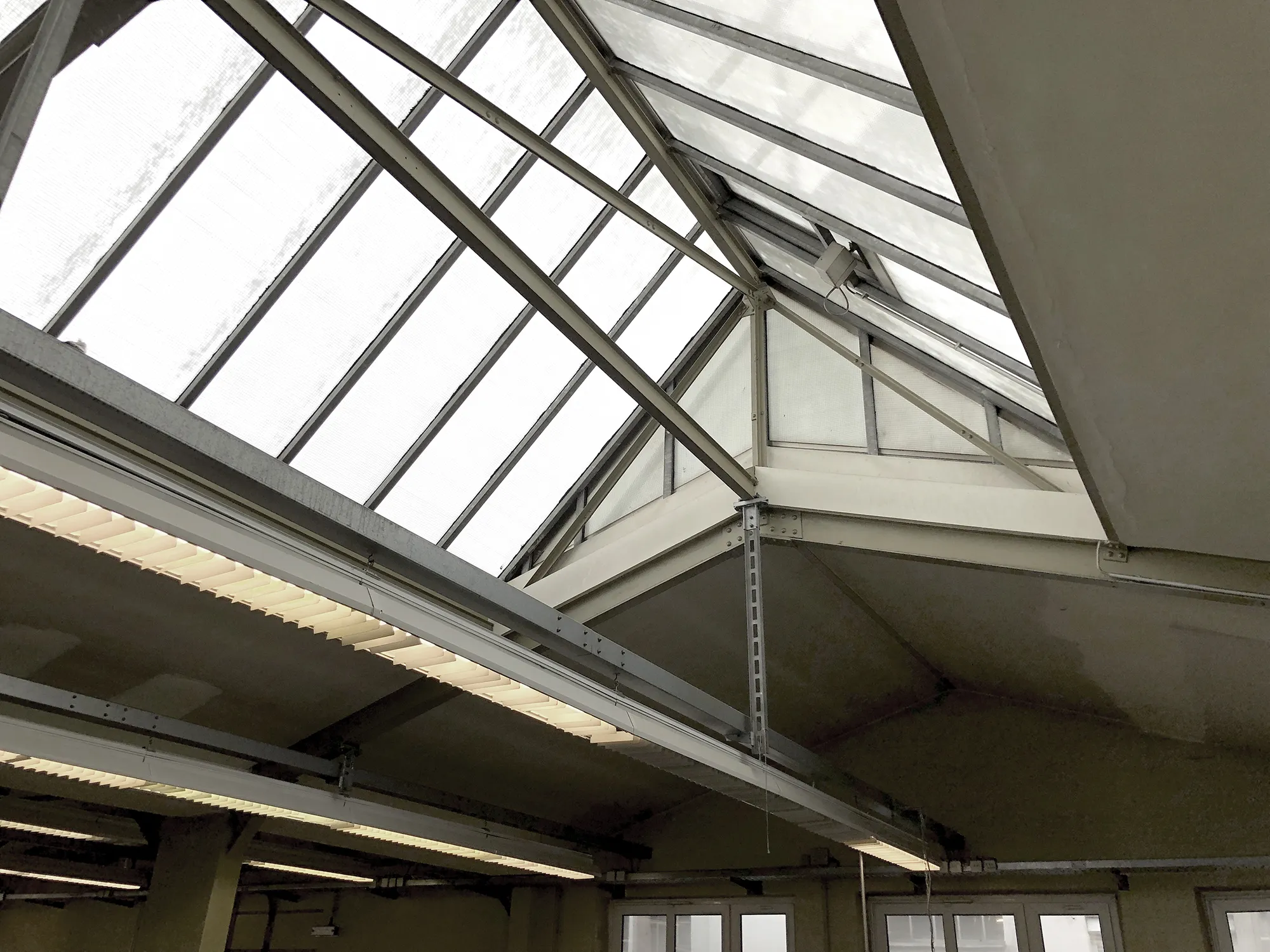 Innenansicht eines Gebäudes mit großem Oberlicht aus Glas und Metallträgern, durch das Tageslicht in den Raum fällt, ergänzt durch Deckenleuchten.