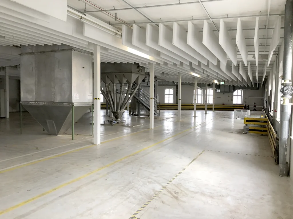 Große Industriehalle mit weißem Boden und Decke, gelb-schwarz markierten Schutzleisten und mehreren großen metallenen Trichtern in der Mitte des Raumes.