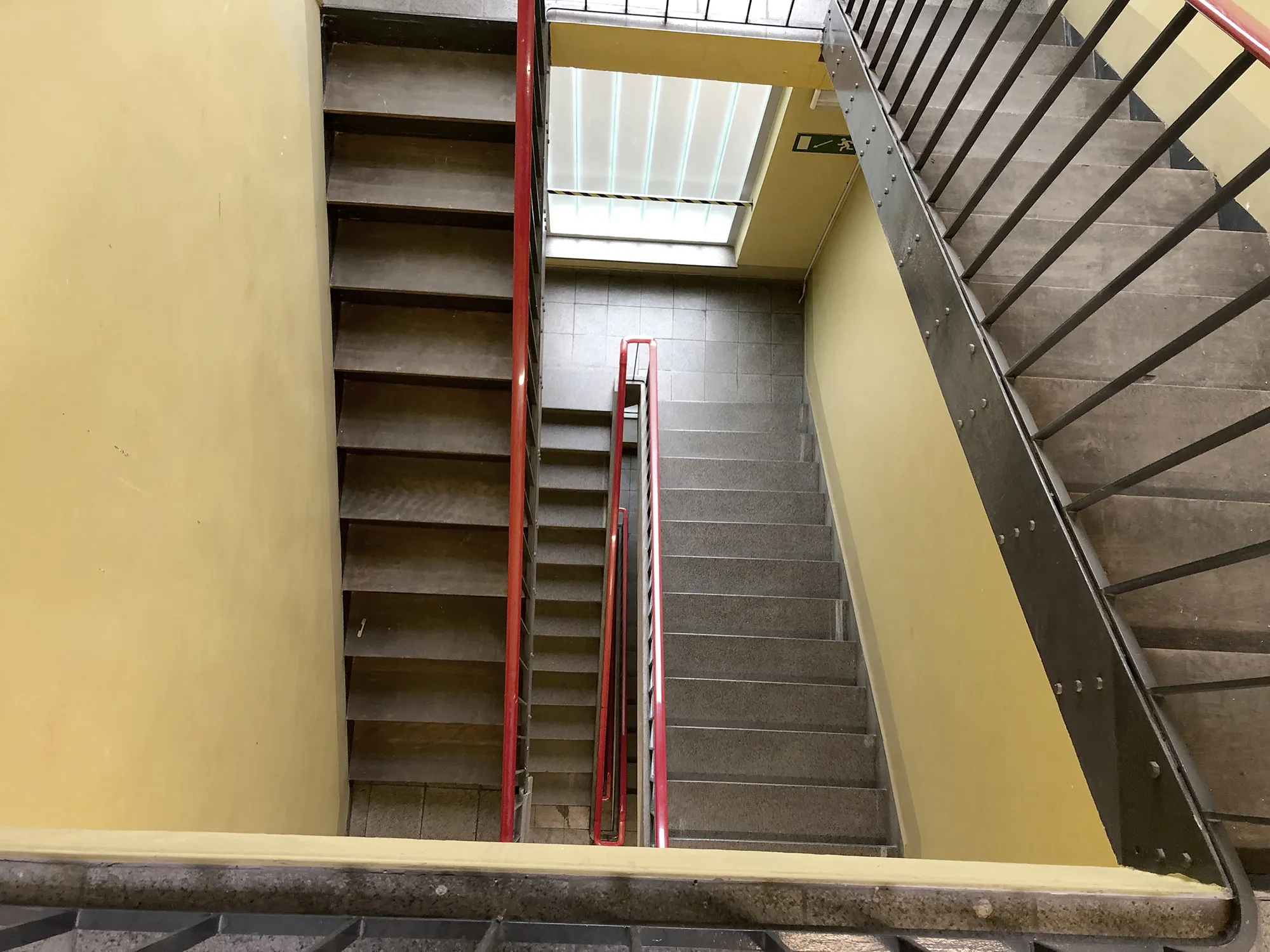 Blick von oben auf ein Treppenhaus mit grauen Stufen, gelben Wänden und roten Handläufen, beleuchtet durch Tageslicht von einem Fenster mit Jalousie.
