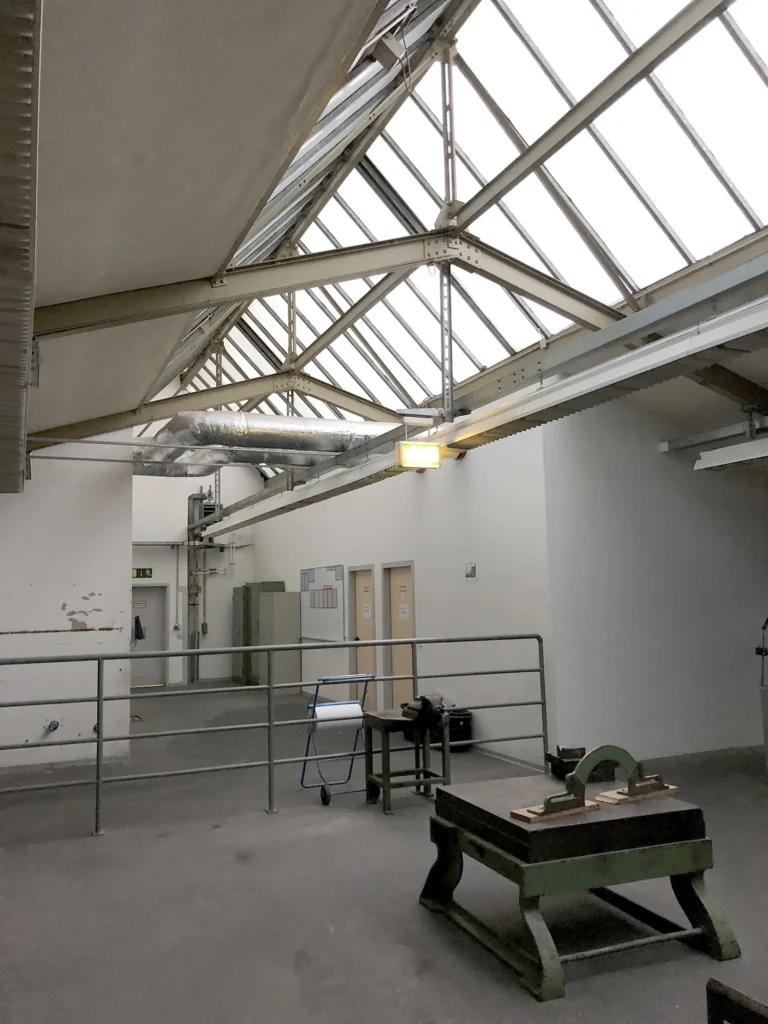 Innenraum einer Industriehalle mit hohem Satteldach aus Glas und Stahlträgern, darunter weiße Wände, mehrere Türen und ein Geländer im Vordergrund, sowie einige Werkbänke und Geräte.