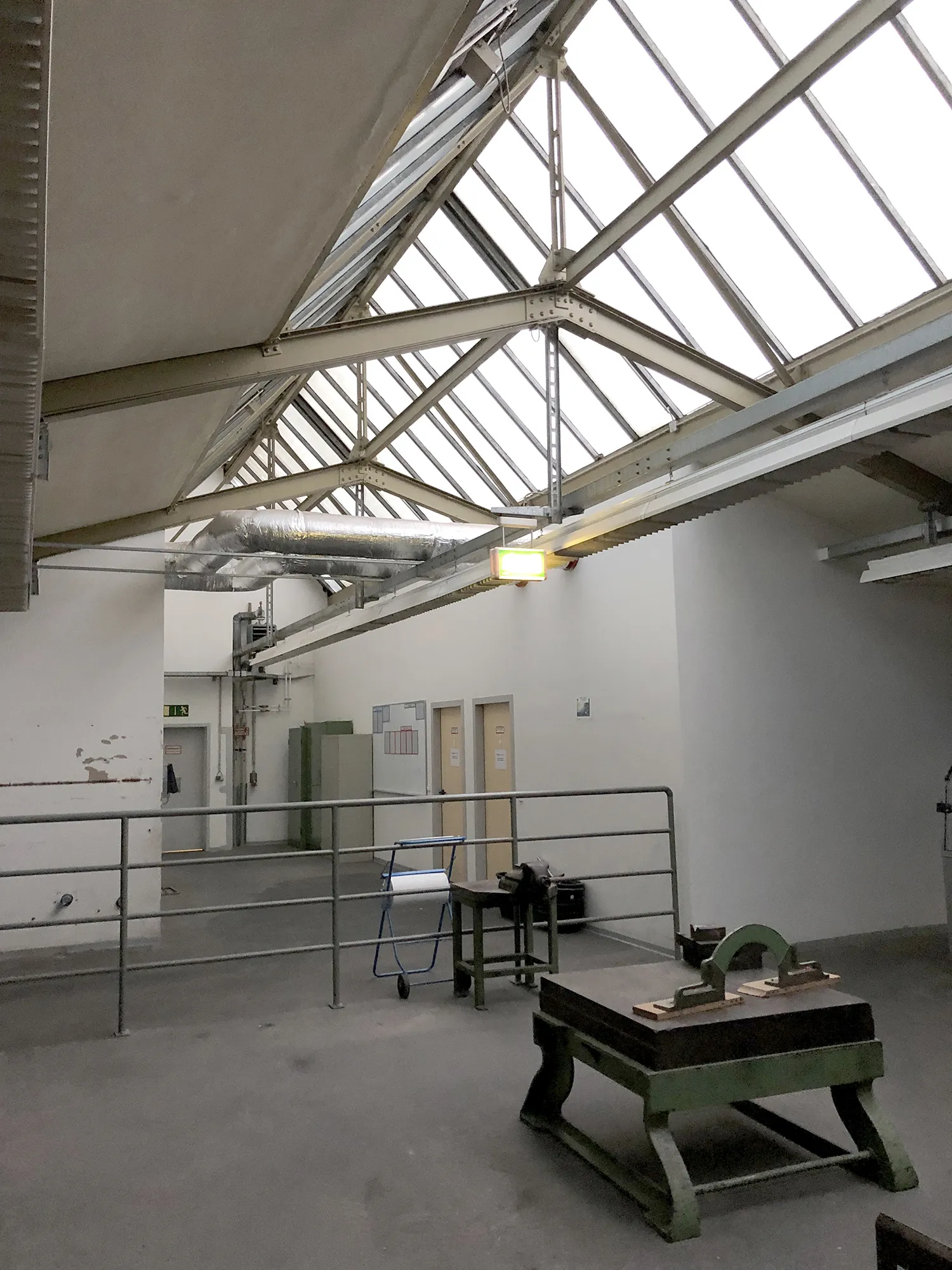 Innenraum einer Industriehalle mit hohem Satteldach aus Glas und Stahlträgern, darunter weiße Wände, mehrere Türen und ein Geländer im Vordergrund, sowie einige Werkbänke und Geräte.