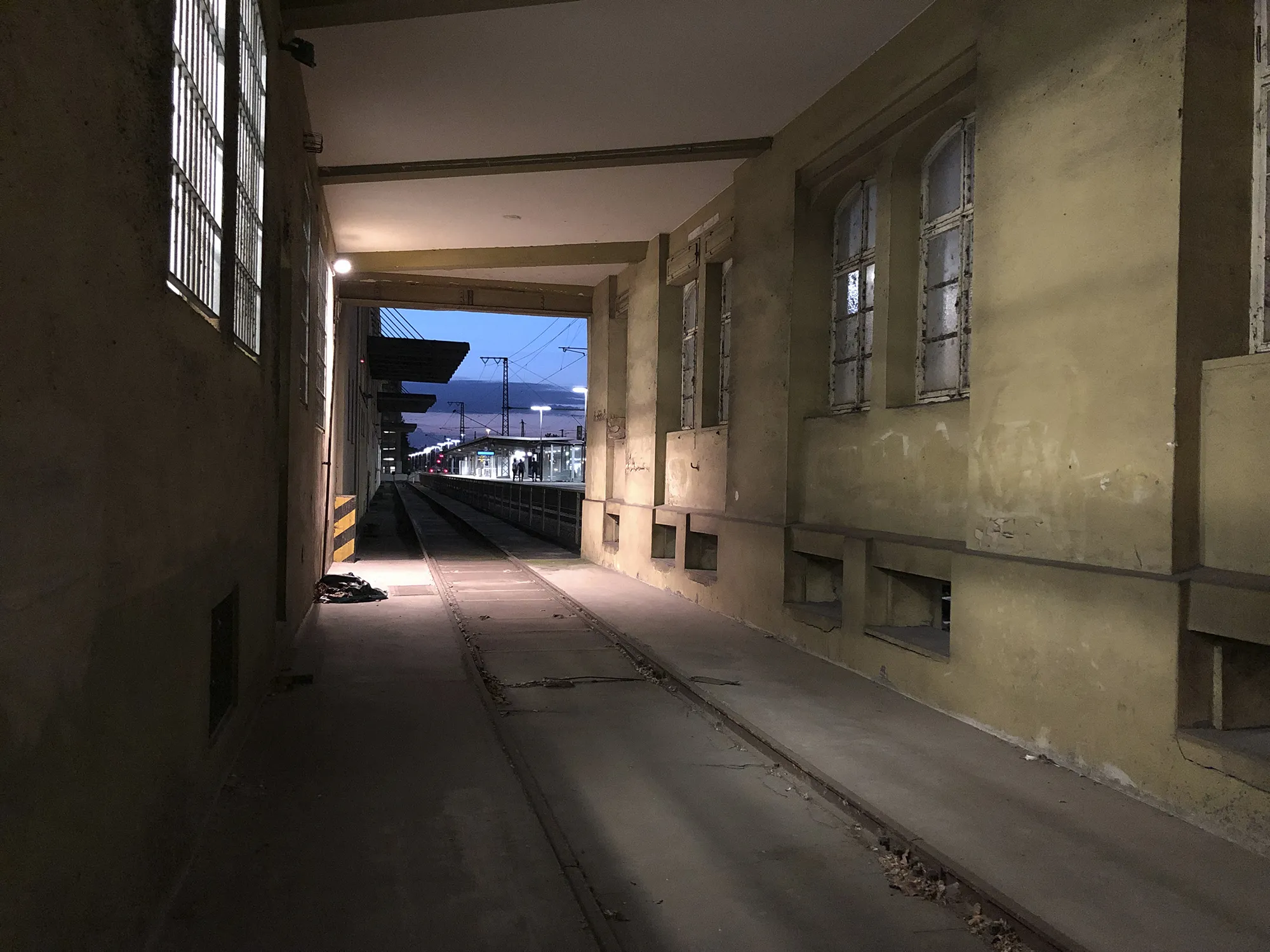Überdachter, schmaler Durchgang mit Schienen im Boden und gelben Wänden, der zu einem beleuchteten Bahnsteig bei Dämmerung führt.