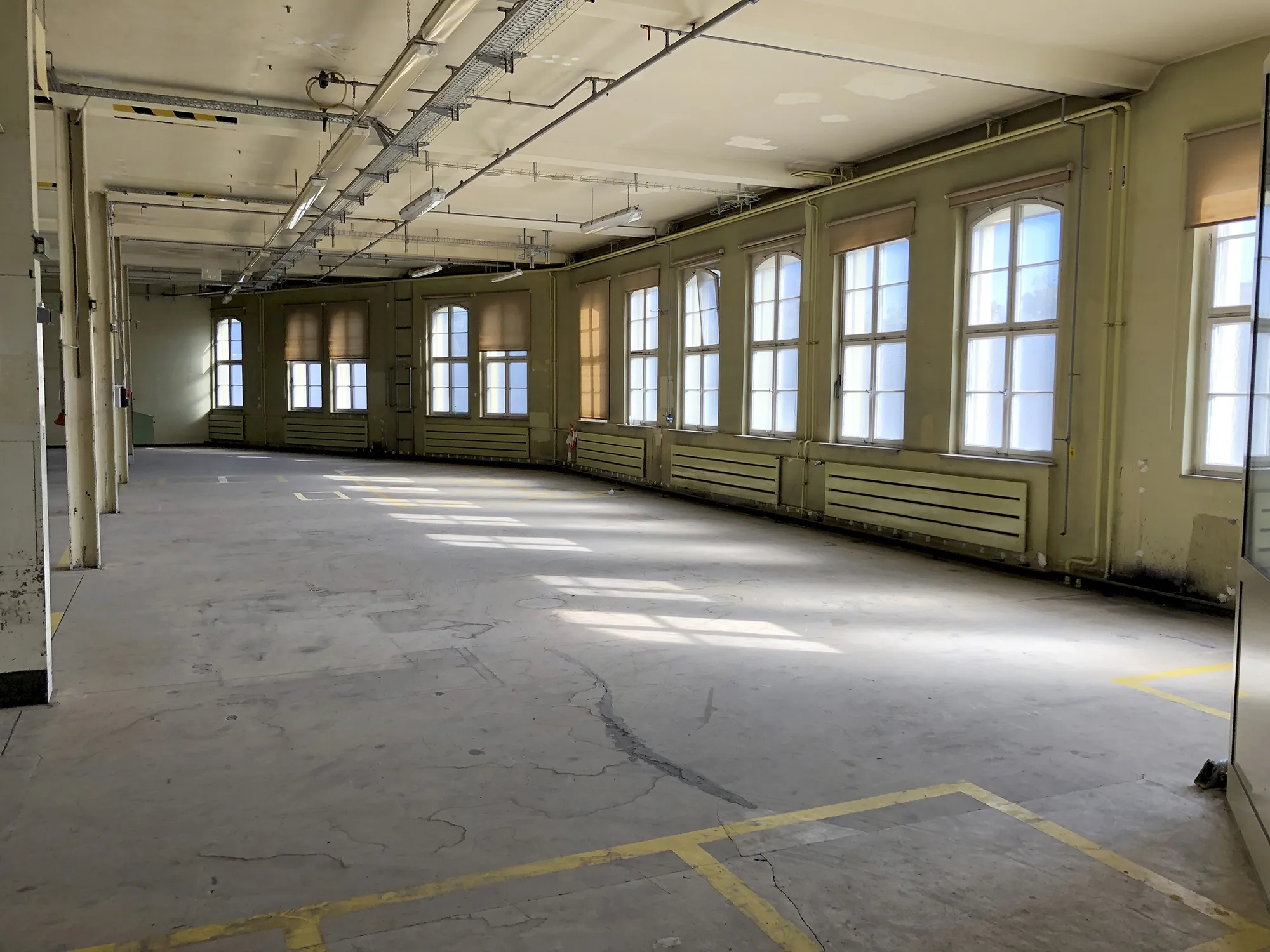 Leere Industriehalle mit gelben Heizkörpern entlang der Fensterwand, mehreren Stützpfeilern und markierten Flächen auf dem Betonboden, beleuchtet durch Tageslicht von großen Fenstern.