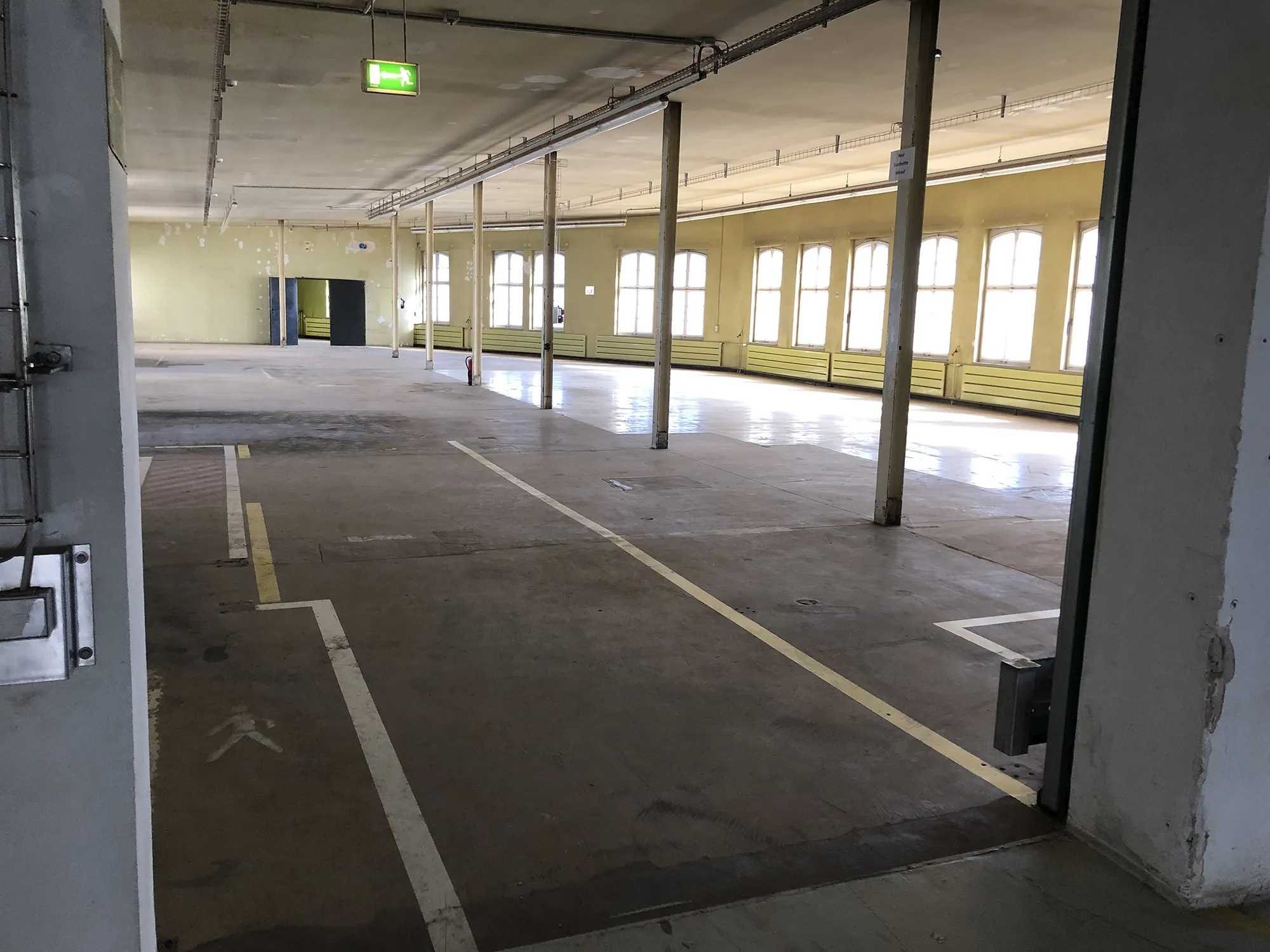 Leere Industriehalle mit gelben Heizkörpern entlang der Fensterwand, mehreren Stützpfeilern und markierten Flächen auf dem Betonboden, beleuchtet durch Tageslicht von großen Fenstern.