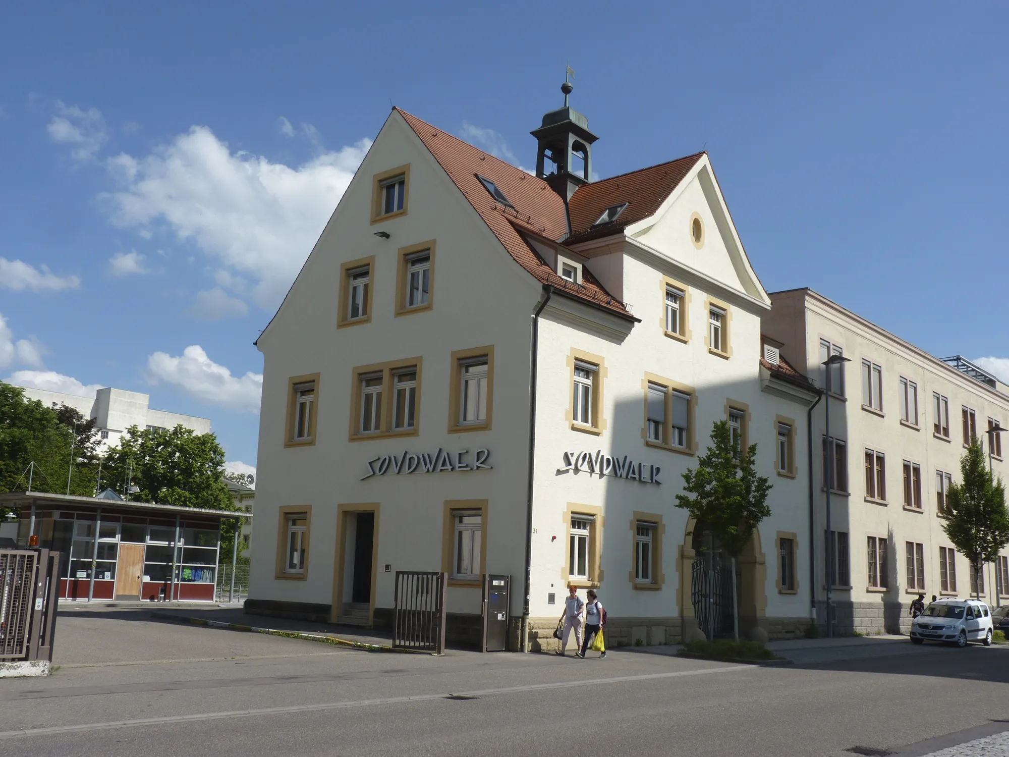 Straßenansicht eines hellen, mehrstöckigen Gebäudes mit rotem Satteldach und kleinem Turm, beschriftet mit dem Schriftzug 'SOVDWAER', bei sonnigem Wetter mit blauem Himmel und vereinzelten Wolken.