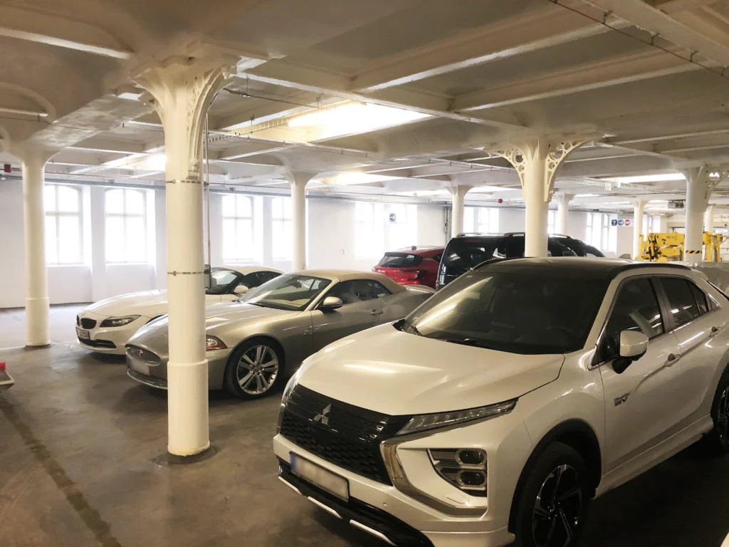 Tiefgarage mit mehreren geparkten Autos. Die Garage hat weiße Stützsäulen mit verzierten Kapitellen und große Fenster, die Tageslicht hereinlassen. Die Decke ist mit sichtbaren Stahlträgern und Beleuchtung ausgestattet.